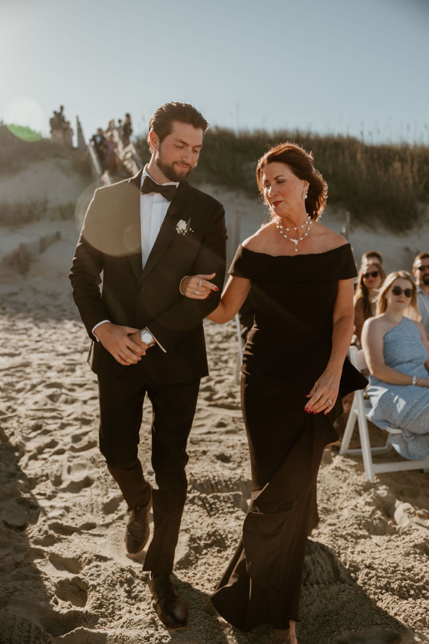 A couple walks together on a sandy beach wearing formal attire during a wedding photoshoot.