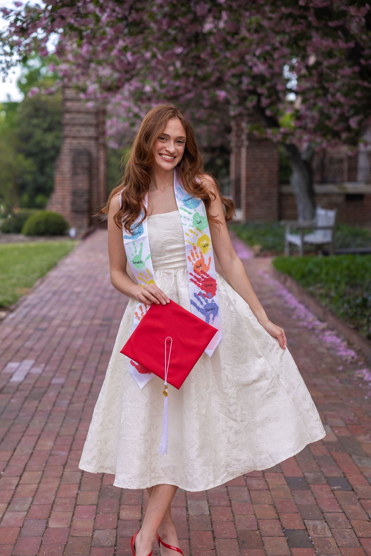 Smiling graduate in white dress holding red cap with colorful sash on brick path with flowering trees.