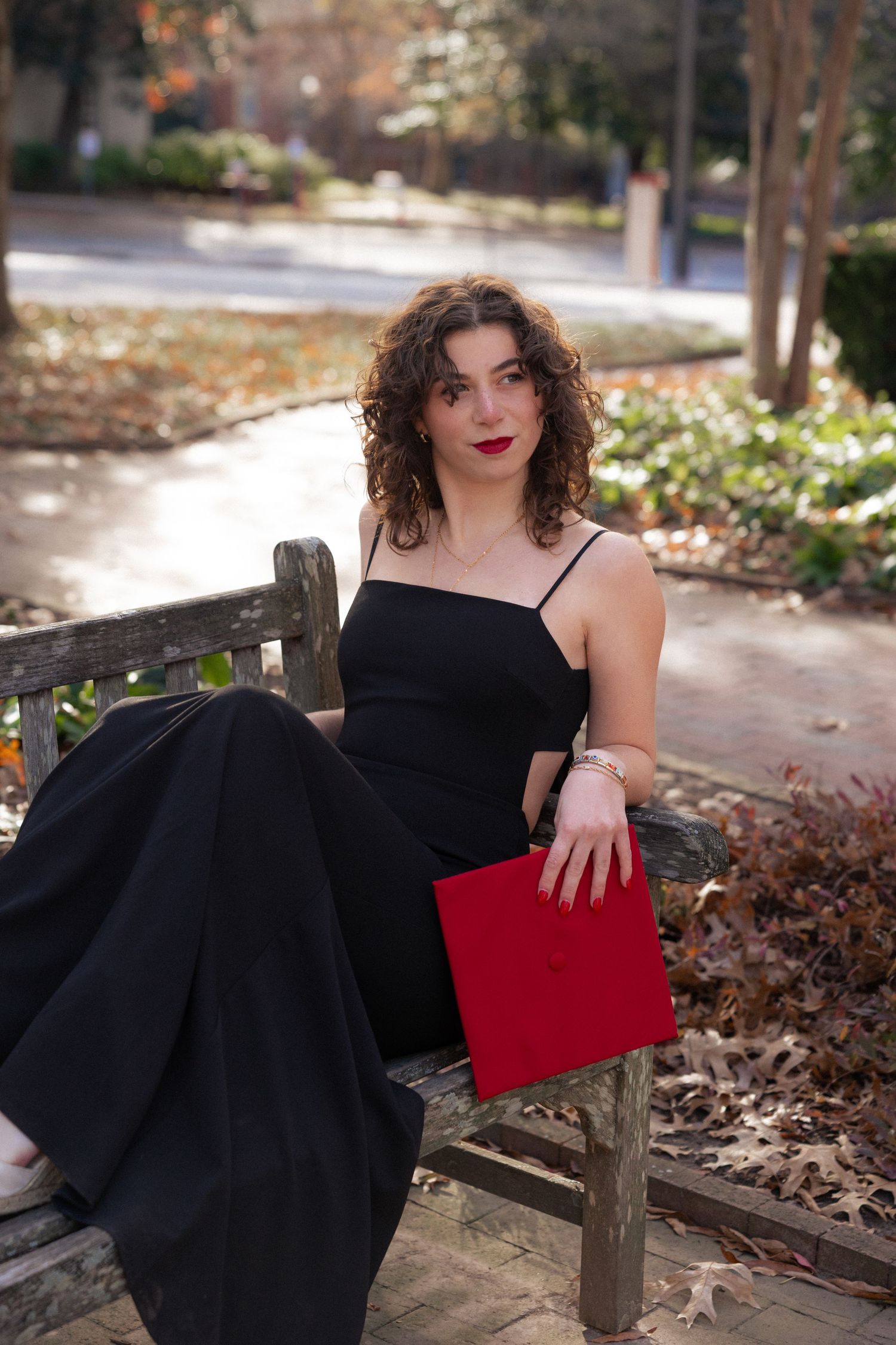 Young woman in black dress holding red book sits on wooden park bench surrounded by autumn leaves.