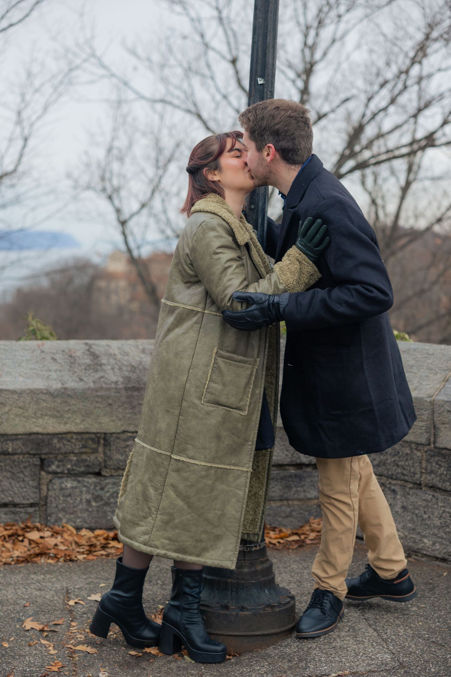 A couple shares a romantic kiss while leaning against a lamppost on a cold autumn day with fallen leaves around them.