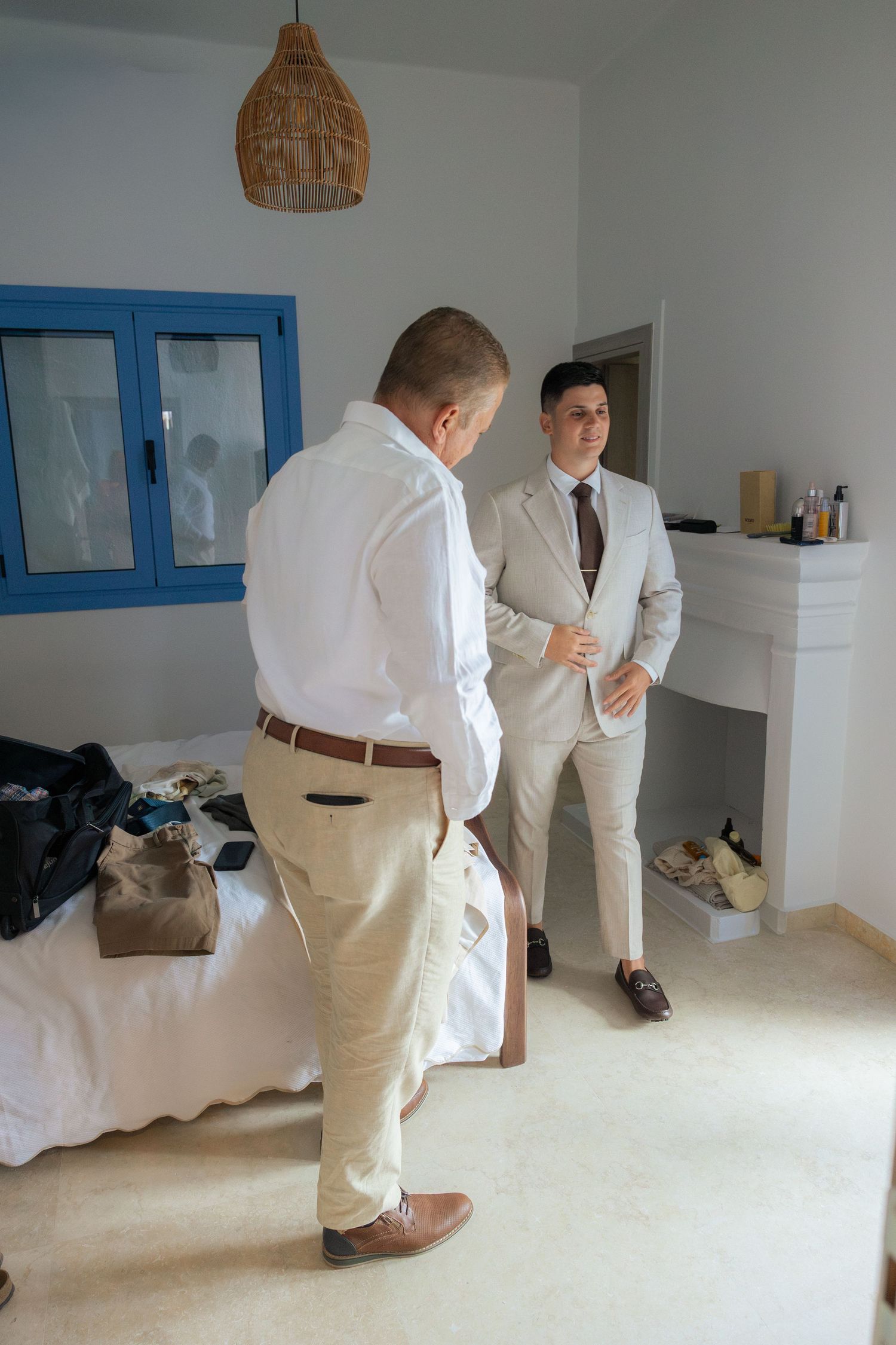 Two people in light colored suits stand in a bright white room with a woven pendant lamp overhead.