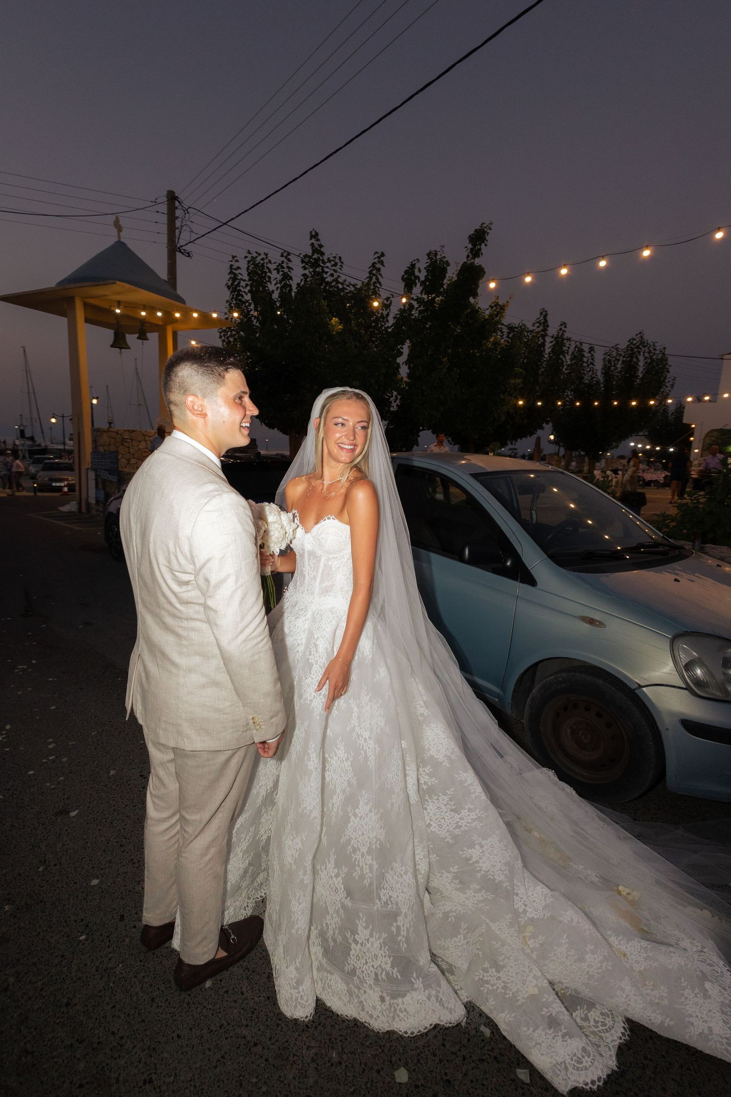 A couple in white wedding attire stands together at dusk under string lights in a parking area.