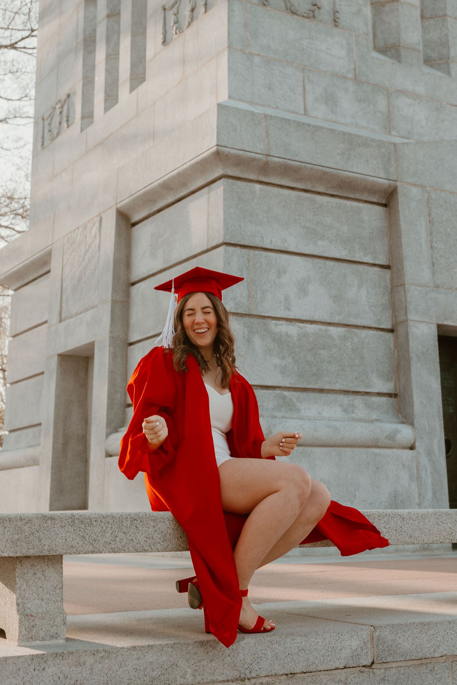 Young woman in red graduation cap and gown celebrating on stone steps outside a university building.