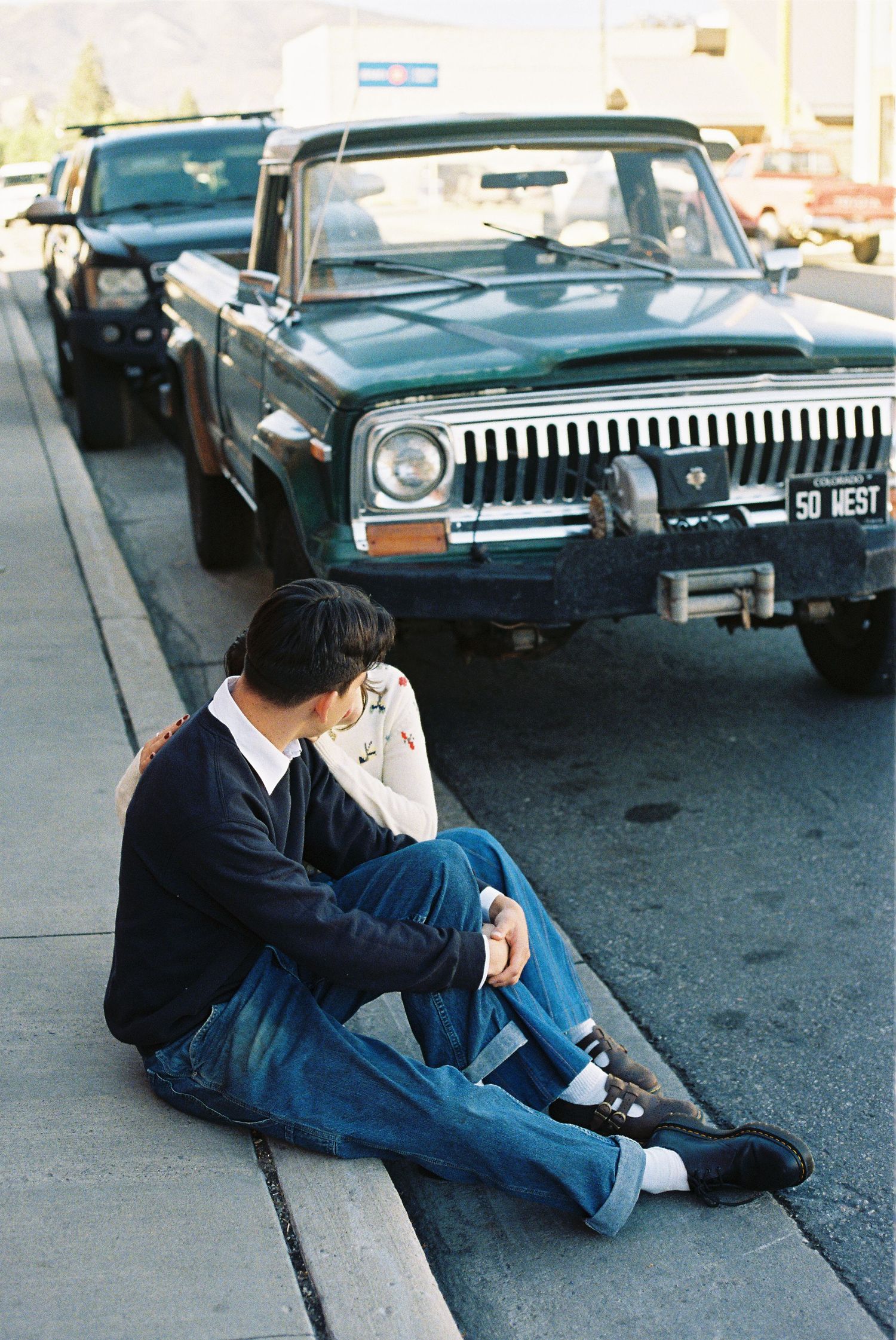 A contemplative person in casual attire sitting on a curb next to a vintage Jeep.