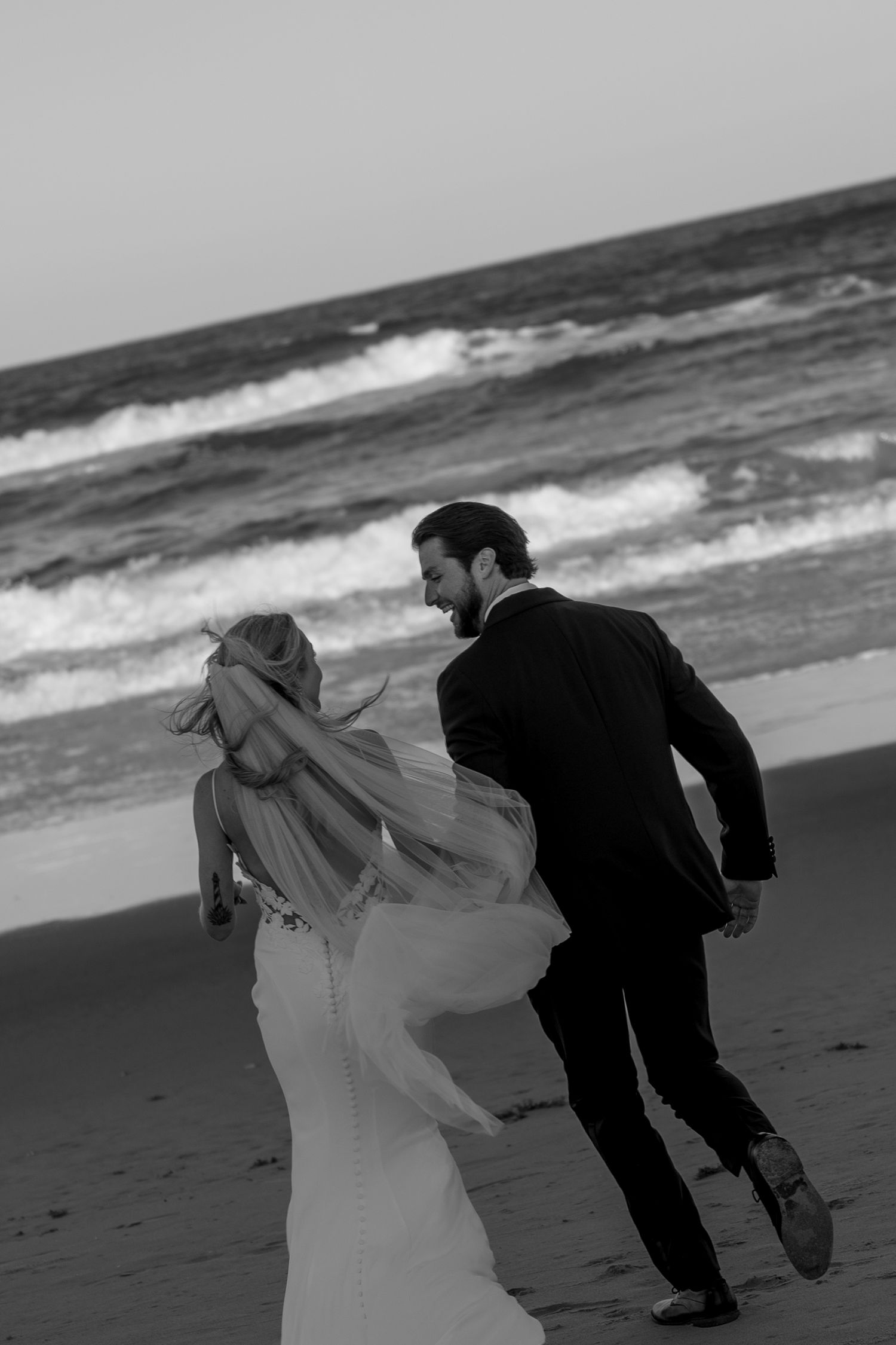 A playful black and white photo of newlyweds running along the beach together.