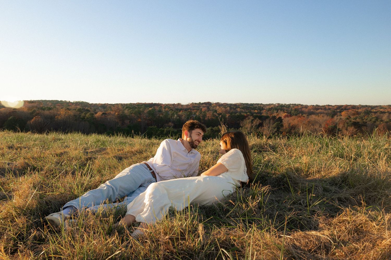 A couple relaxes together in a grassy field during golden hour with rolling hills in the background.