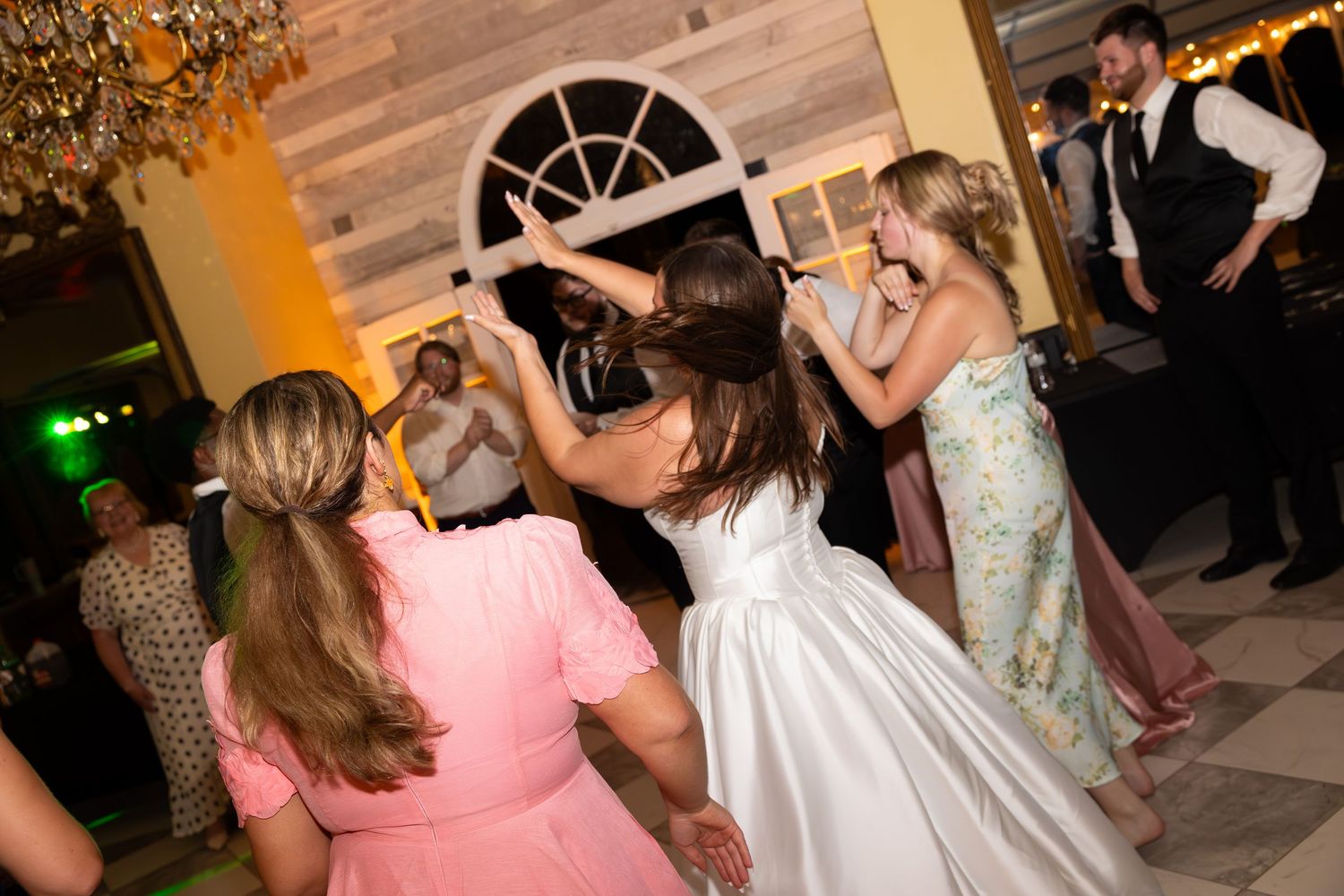 Wedding reception guests dancing and celebrating on a wooden dance floor with festive lighting.