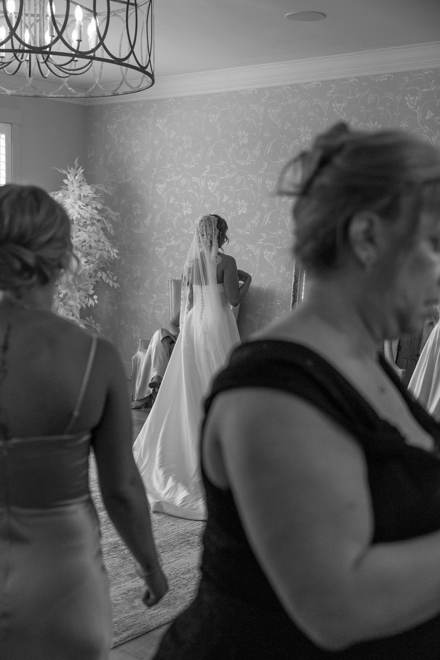 Black and white photo of a bride getting ready in a room with bridesmaids and an ornate chandelier.