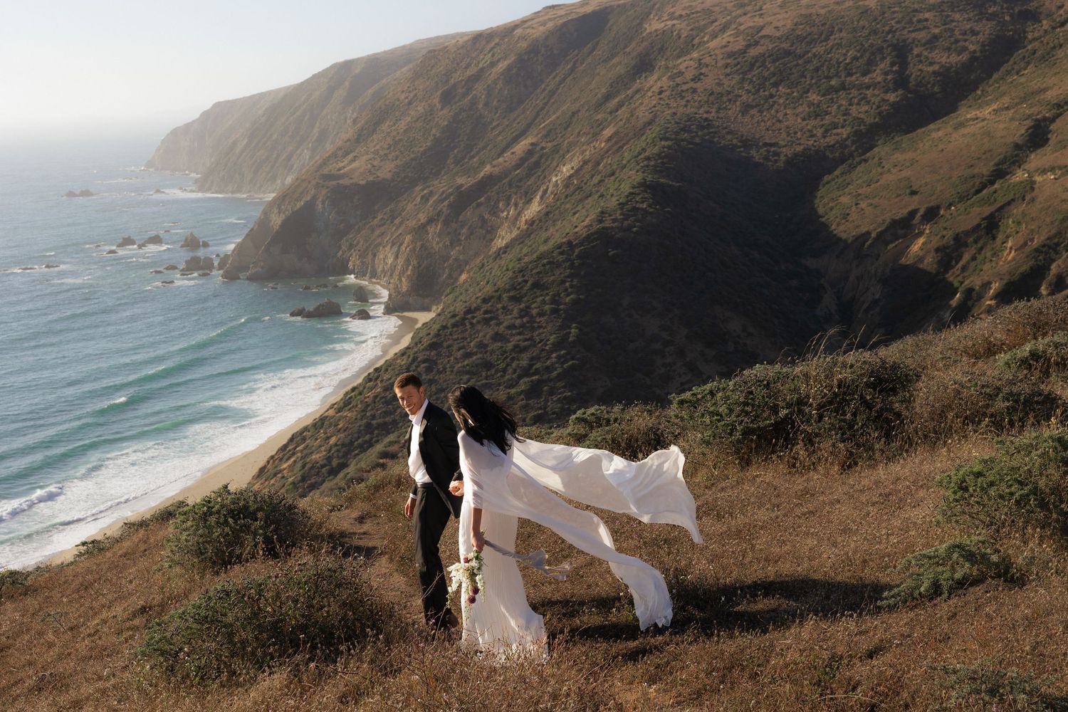 A couple in wedding attire walks along a scenic coastal cliff overlooking the Pacific Ocean on a cloudy day.