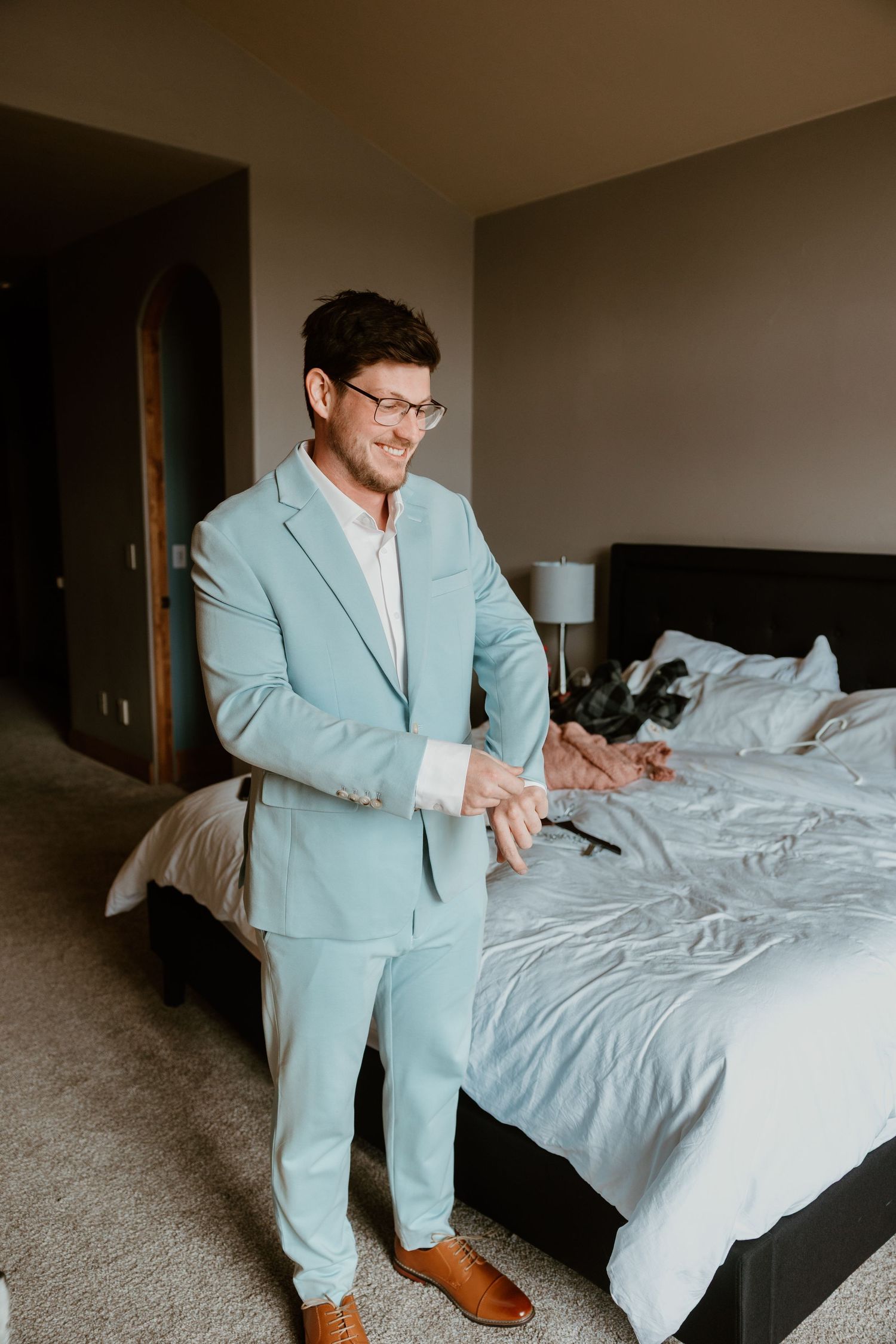 A person in a light blue suit stands in a bedroom looking at their wedding attire.