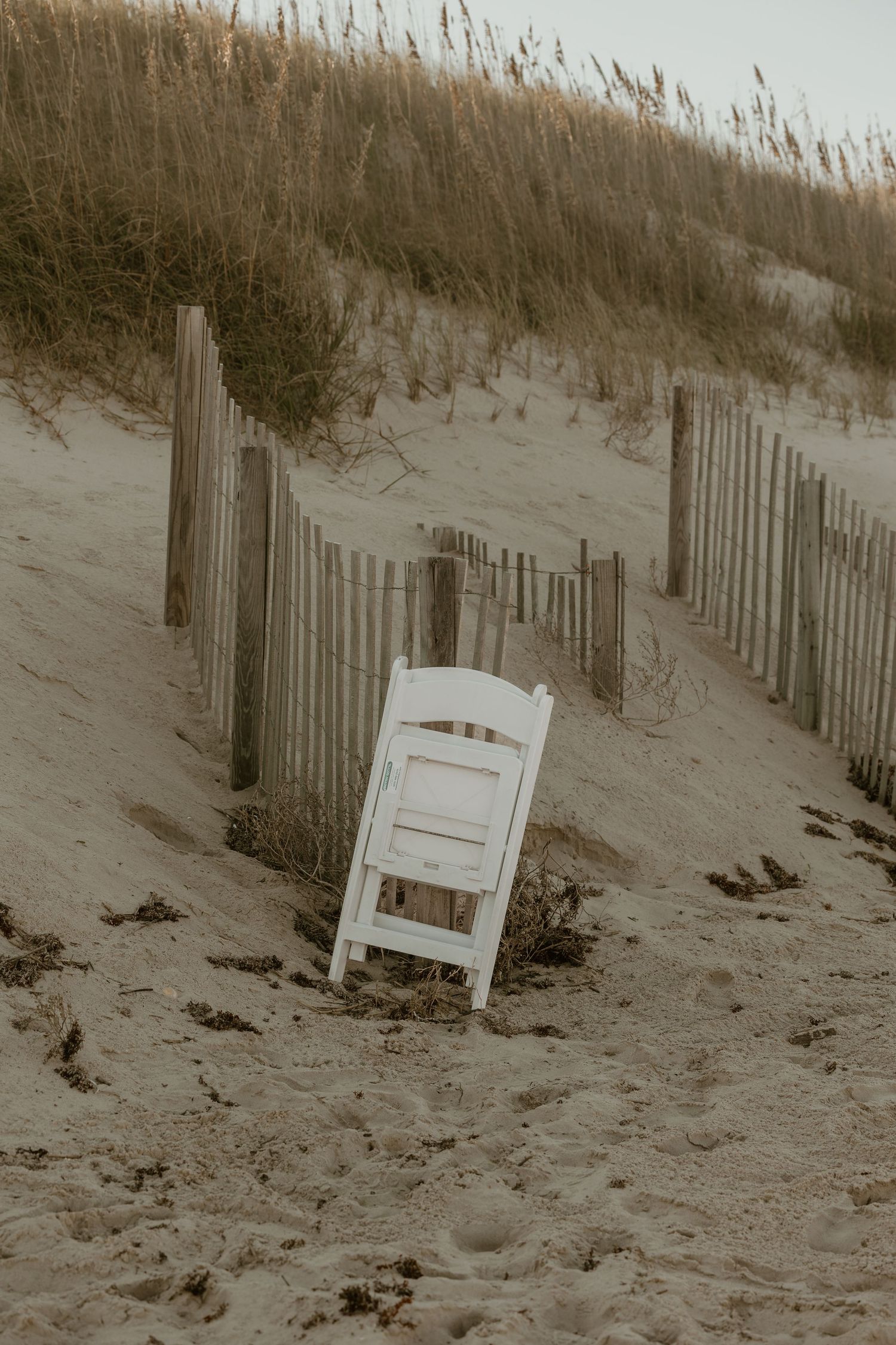 Wooden beach chair sits alone among sand dunes and sea grass fencing.