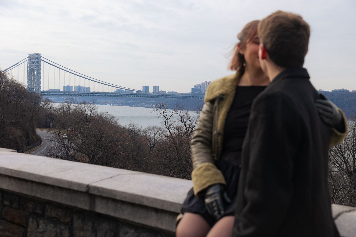 Two people enjoy a scenic overlook with a bridge and city skyline visible in the distance.