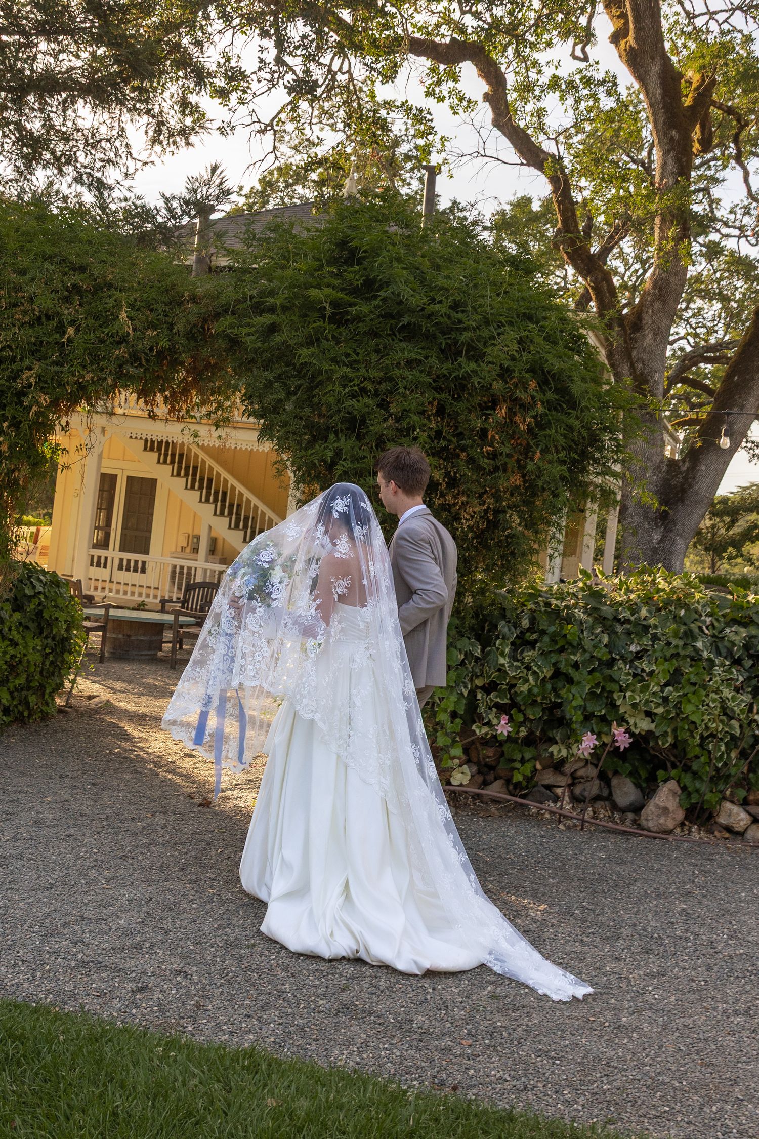 A bride in a flowing white gown and veil walks along a gravel path beside a yellow building in an outdoor garden setting.
