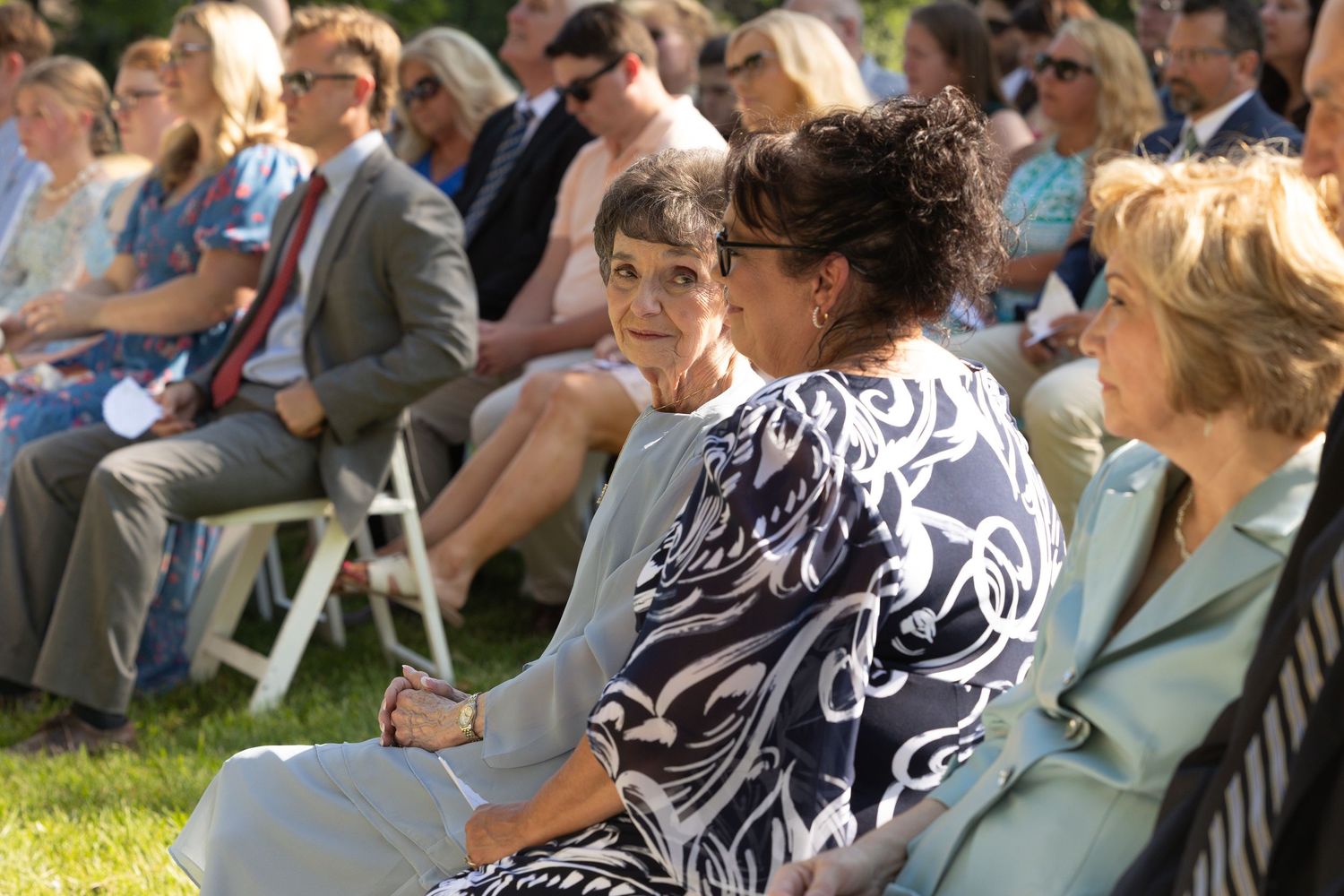 Wedding guests seated in white chairs on a lawn during an outdoor ceremony.