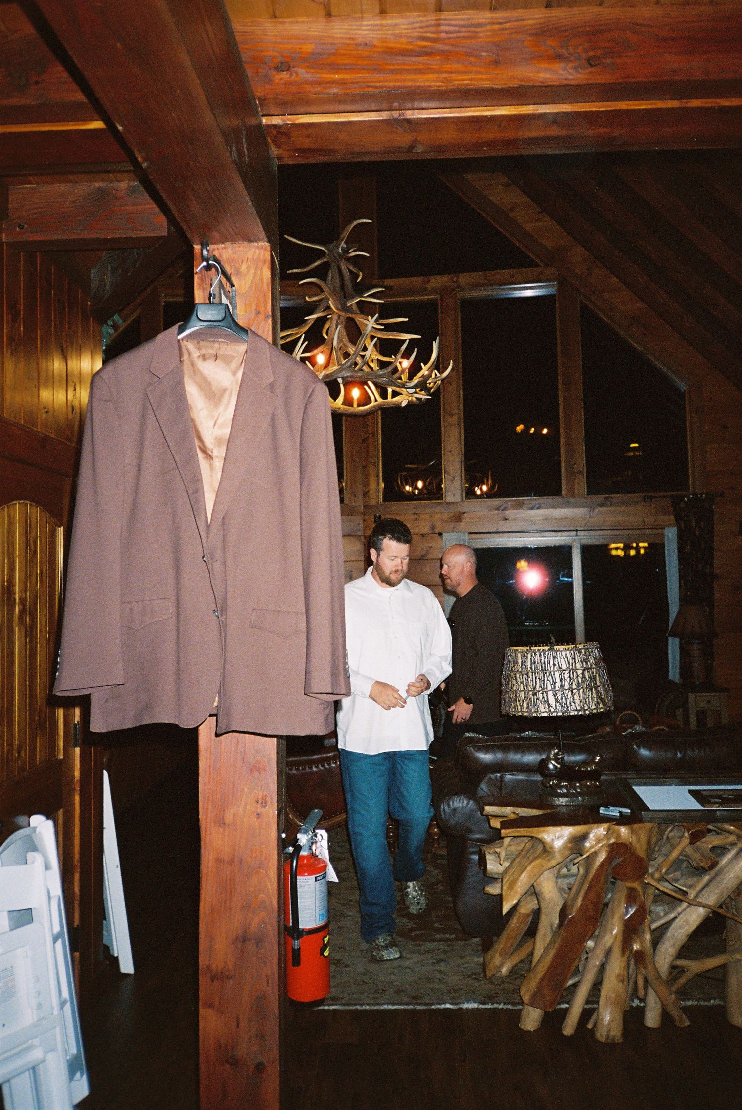 Rustic lodge interior with wooden beams, antler chandelier, and casual gathering in a dimly lit room.