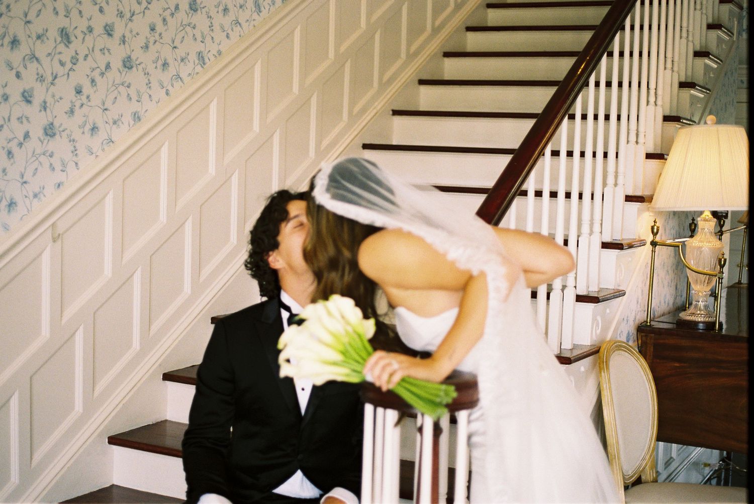 Wedding couple dances and poses on an elegant white staircase with floral wallpaper.