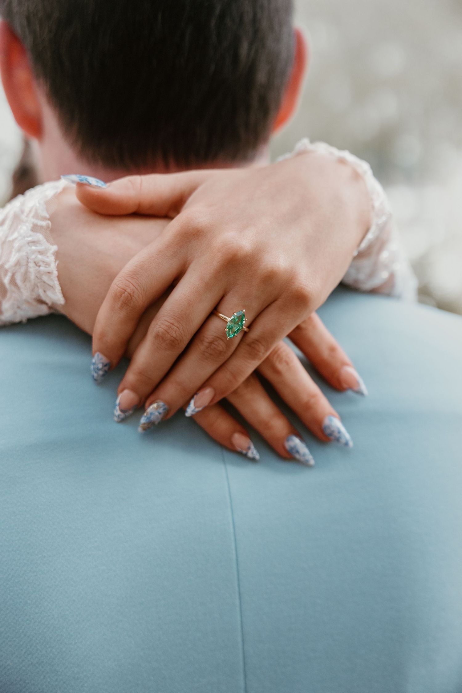 Close-up of hands wearing an emerald ring and sparkly nail polish against a light blue background.