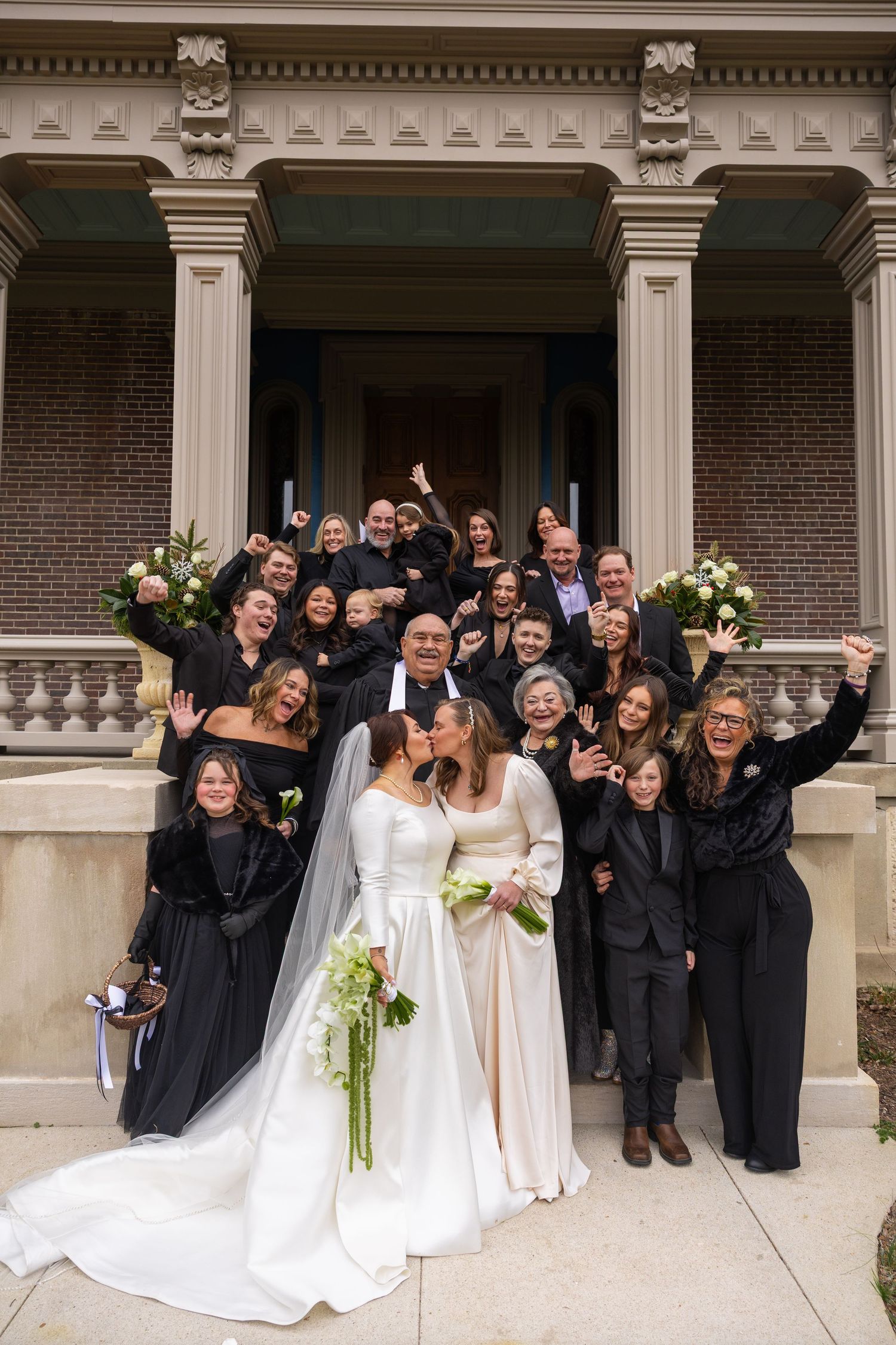 Wedding party poses on grand stone steps of brick building with ornate columns and white floral arrangements.