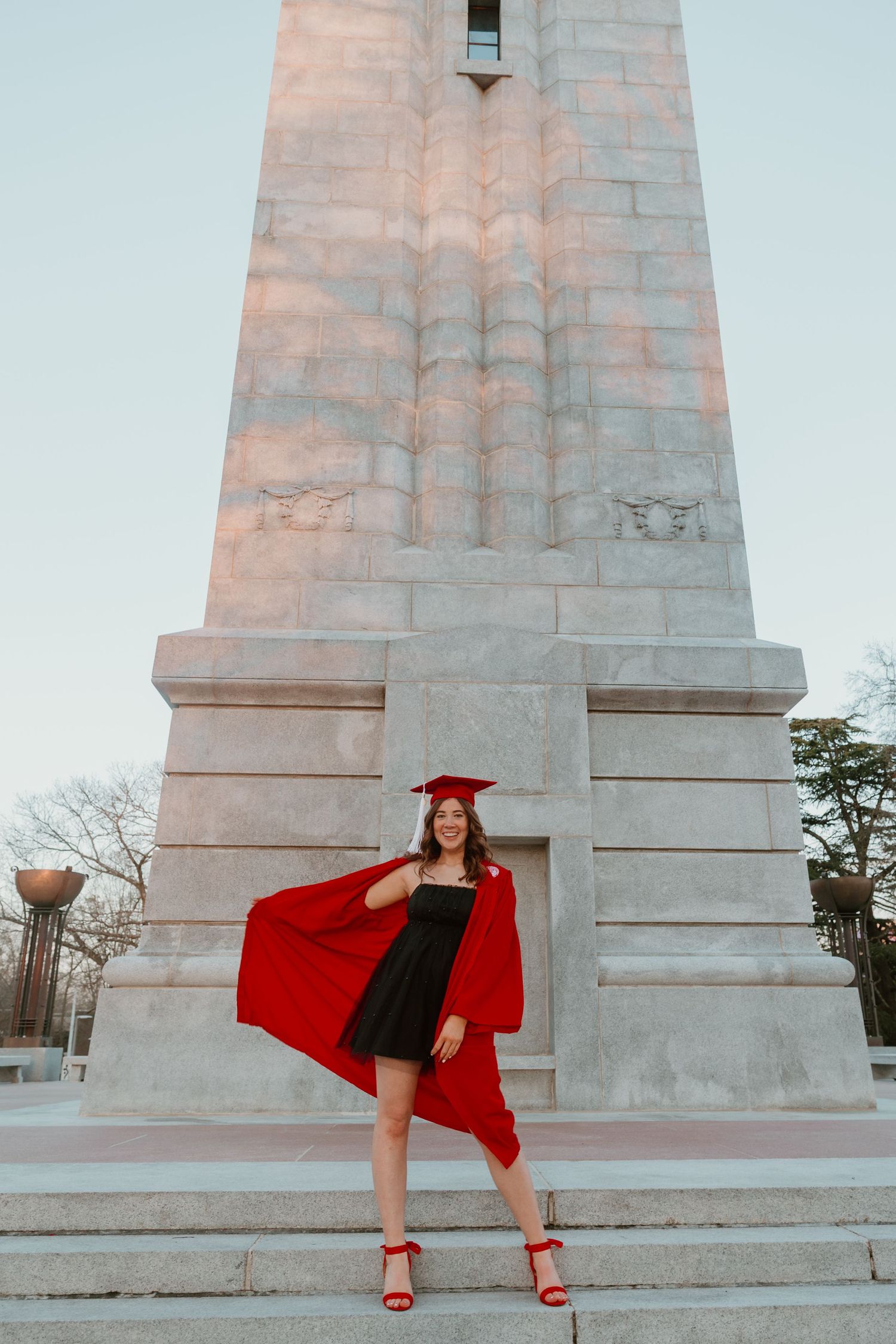 A smiling graduate in red cap and gown poses in front of a tall stone monument, wearing a black dress and red heels.