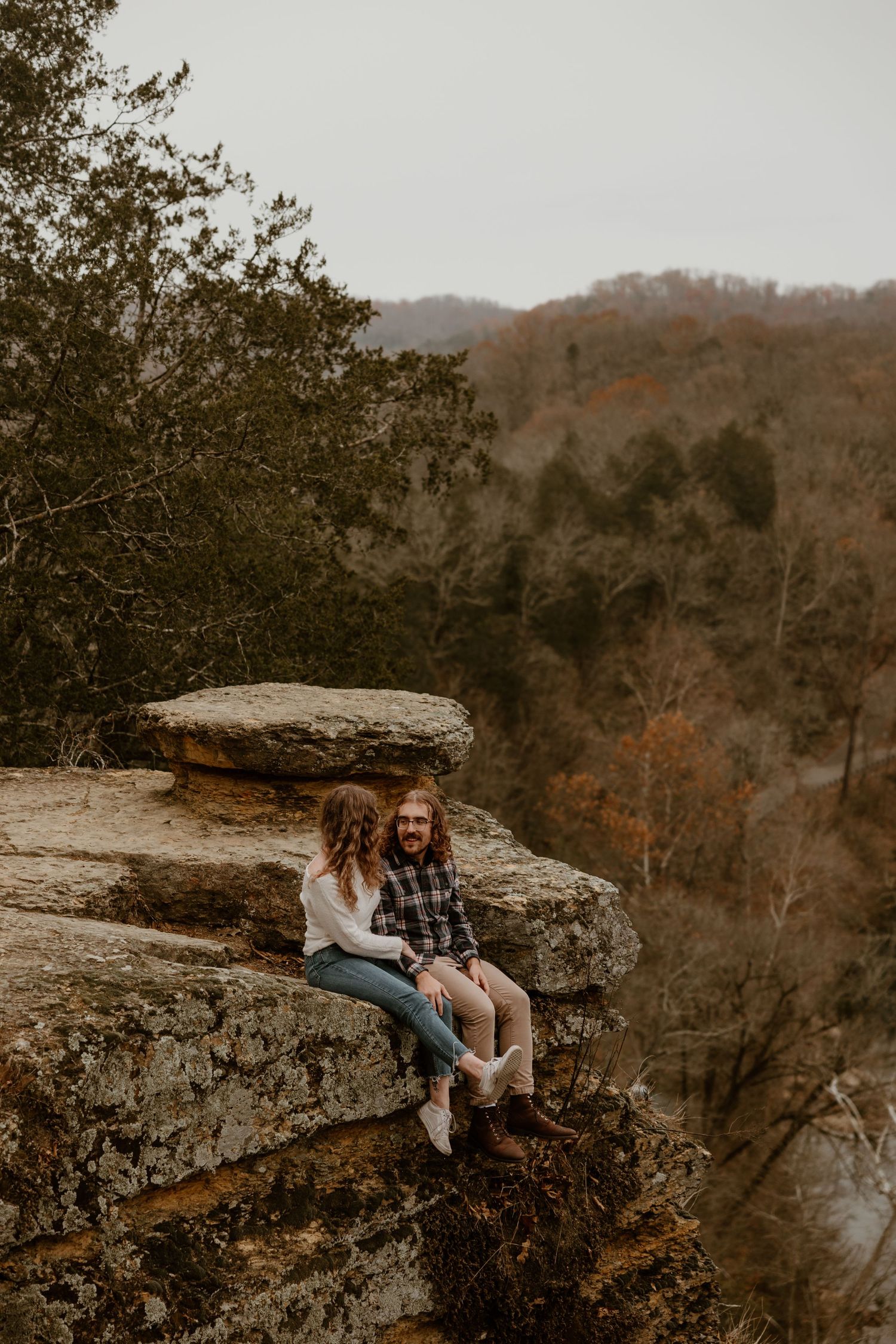 Two people sitting on edge of rocky overlook with autumn mountain landscape behind them.