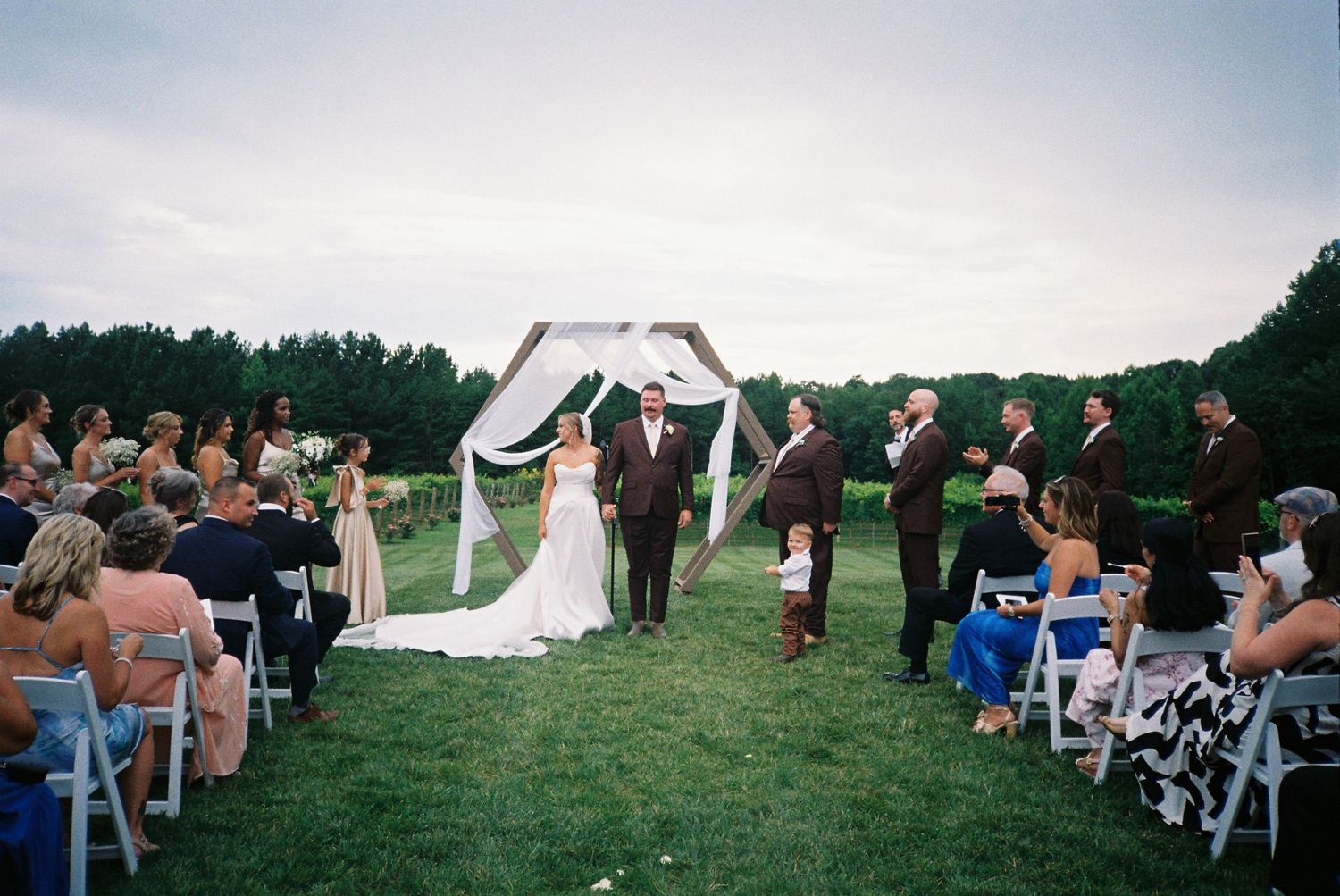 Wedding guests gather outdoors on a grassy hillside with white chairs arranged in rows for a scenic ceremony.