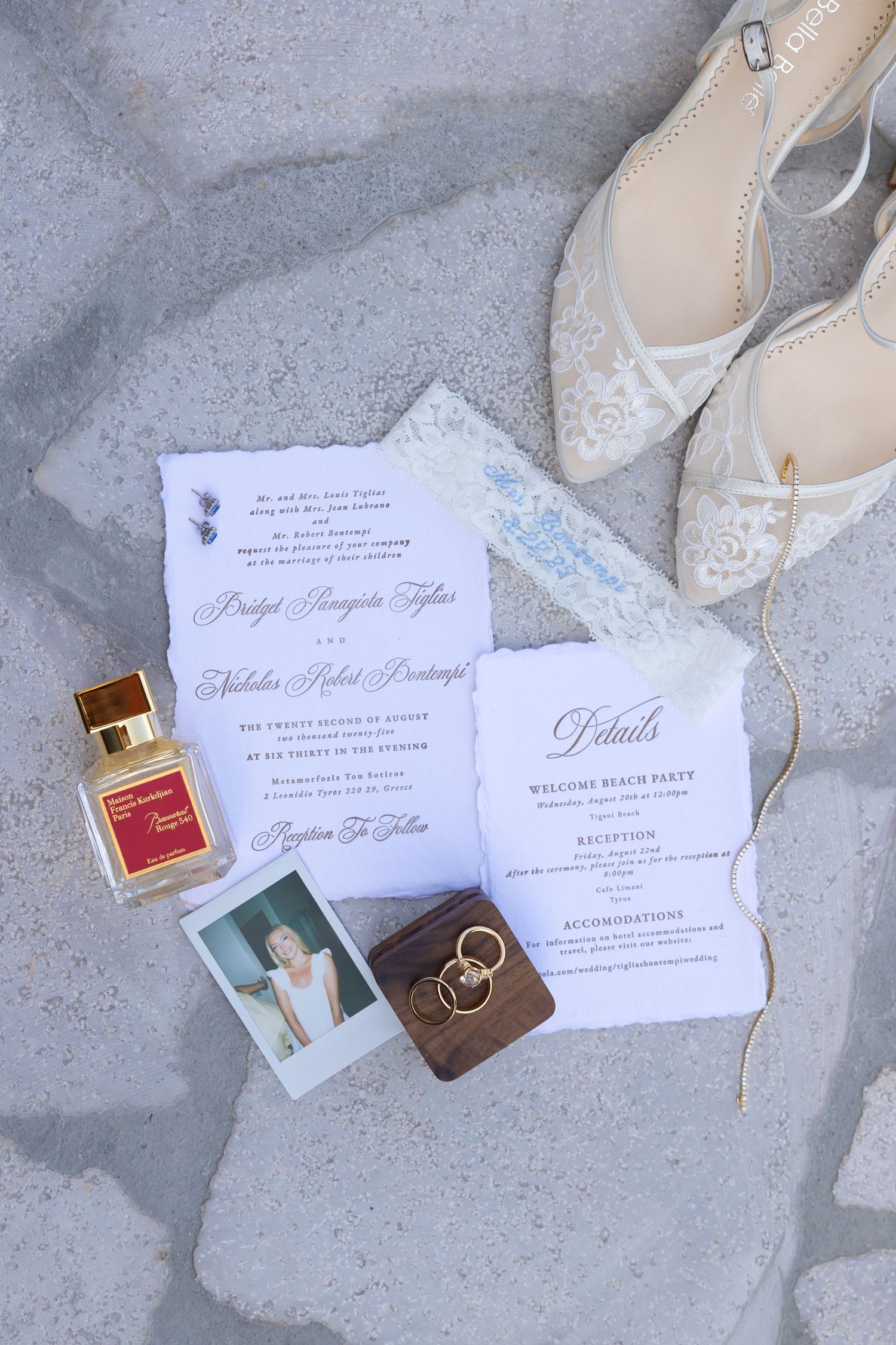 Wedding details flatlay with invitations, perfume bottle, rings and delicate lace shoes on light gray surface.