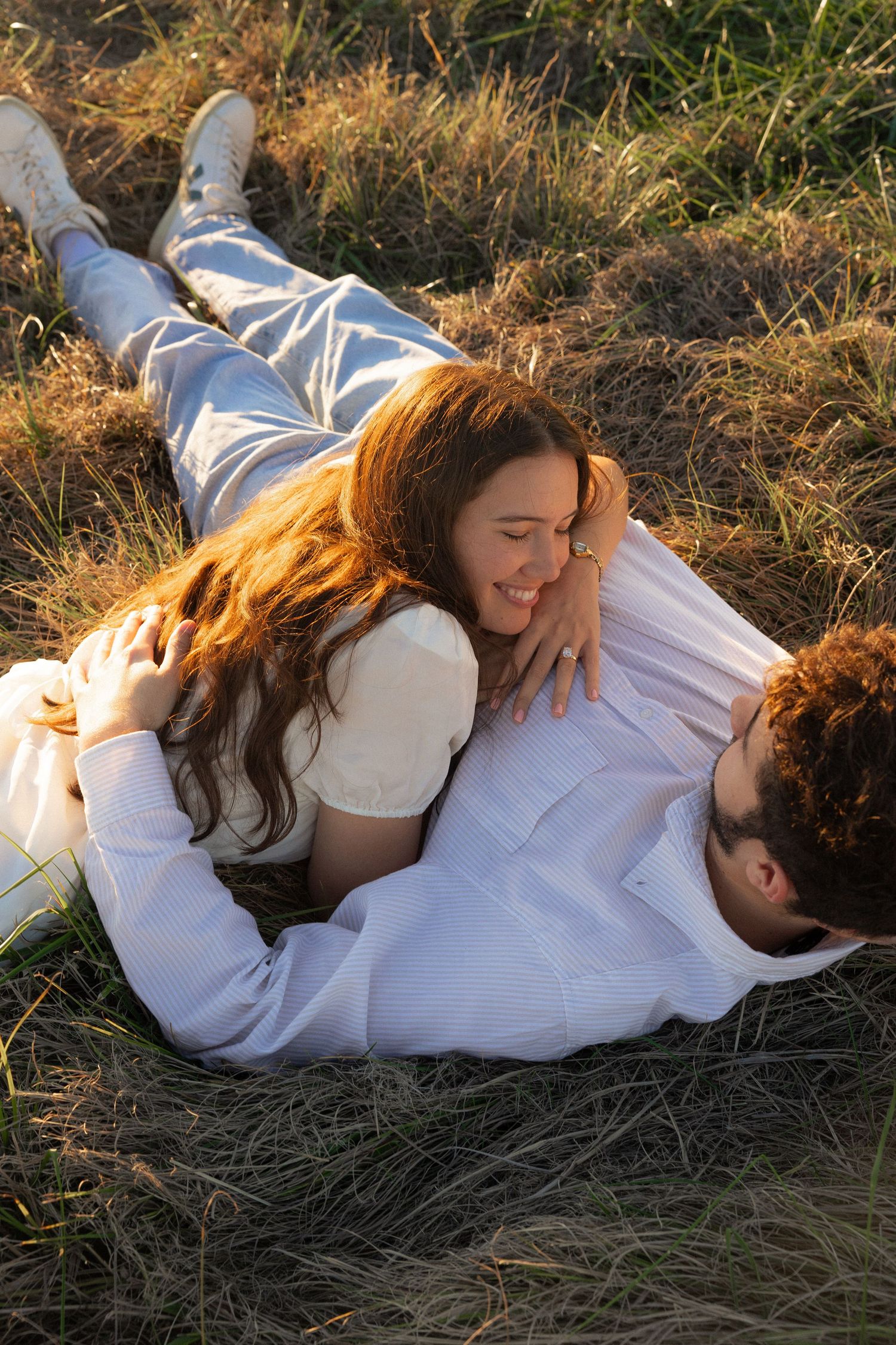 A couple in white clothing lies together in golden grass during sunset.