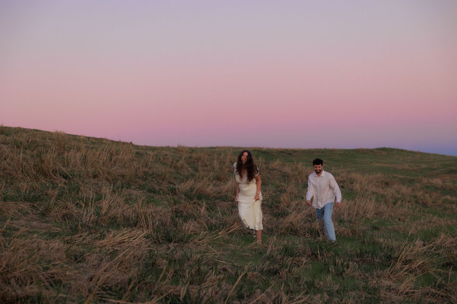 A couple walks through a golden field at sunset creating a romantic silhouette against the pink sky.