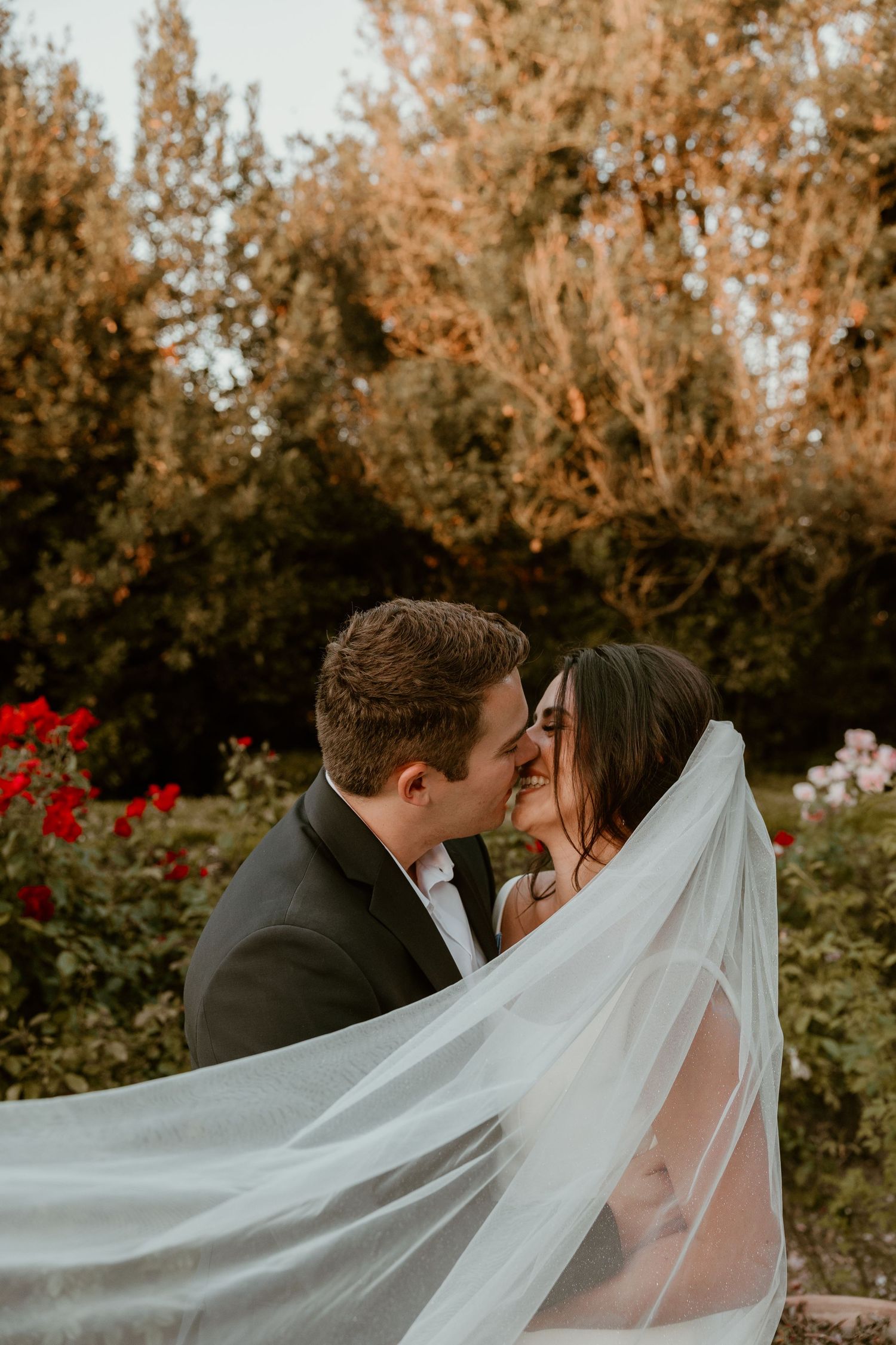 Romantic wedding couple shares intimate kiss under flowing veil in garden setting.