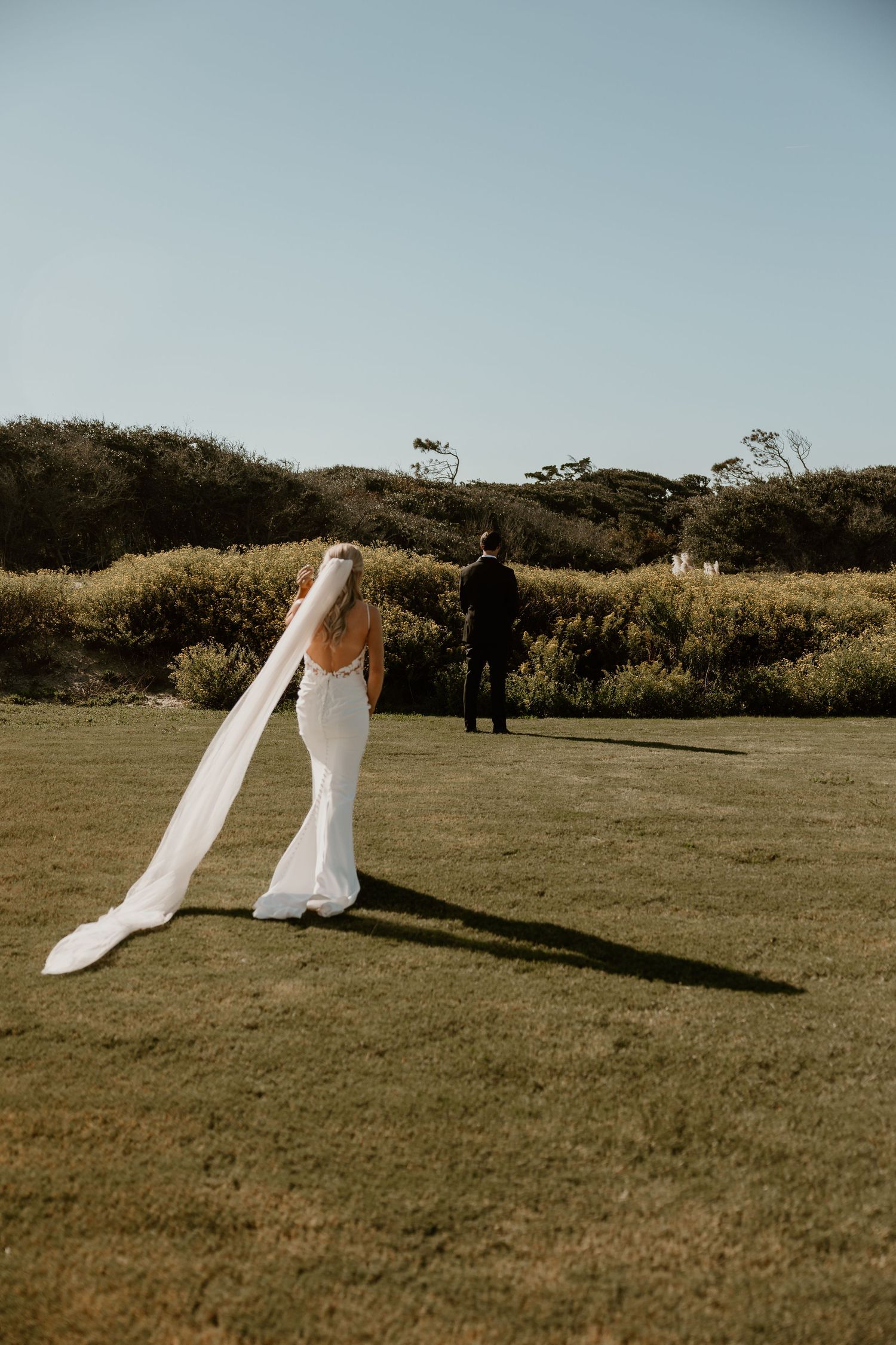 Bride in flowing white dress with veil standing on grassy coastal hilltop.