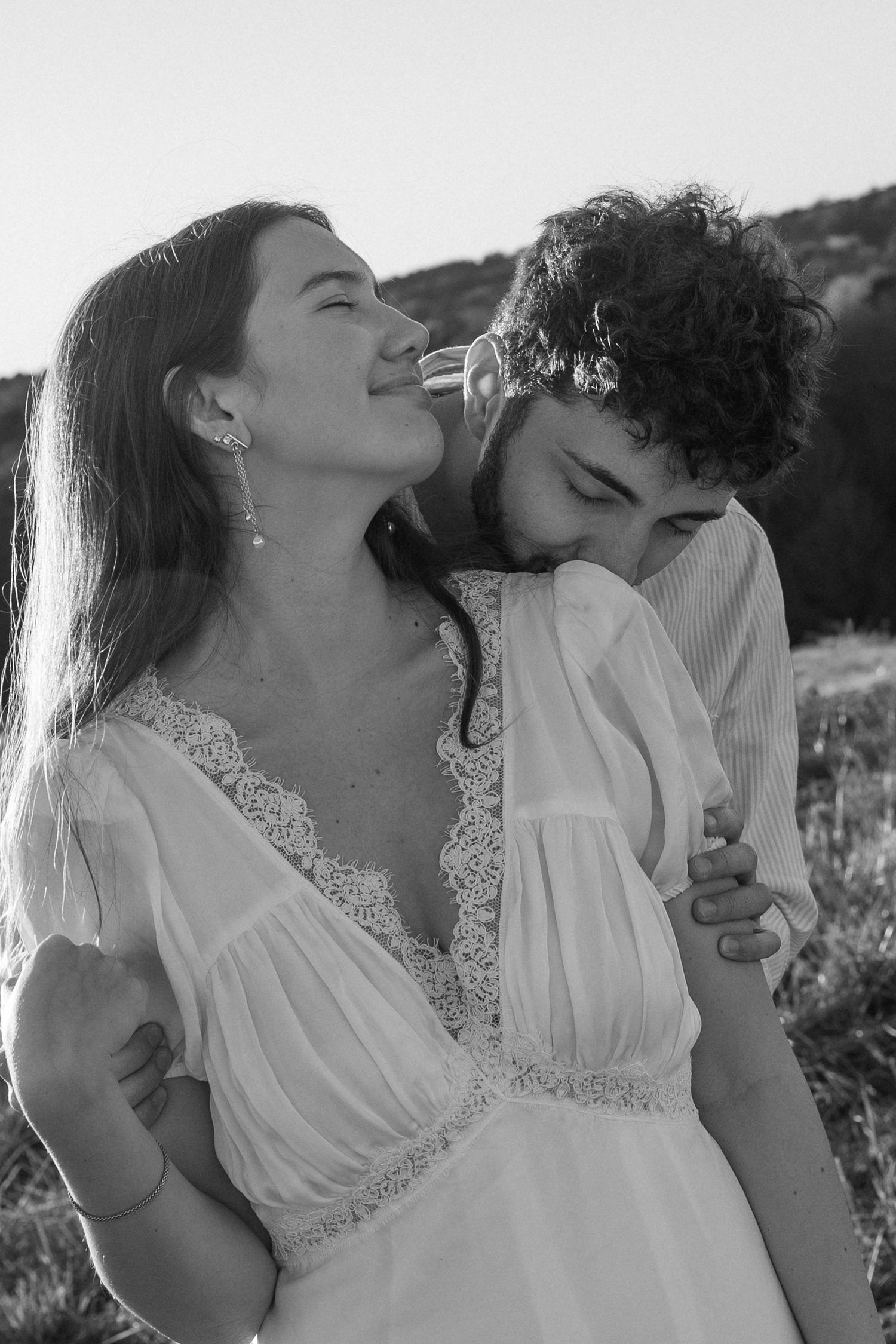 A couple in white attire share a drink from wine bottles in a romantic outdoor photoshoot series.