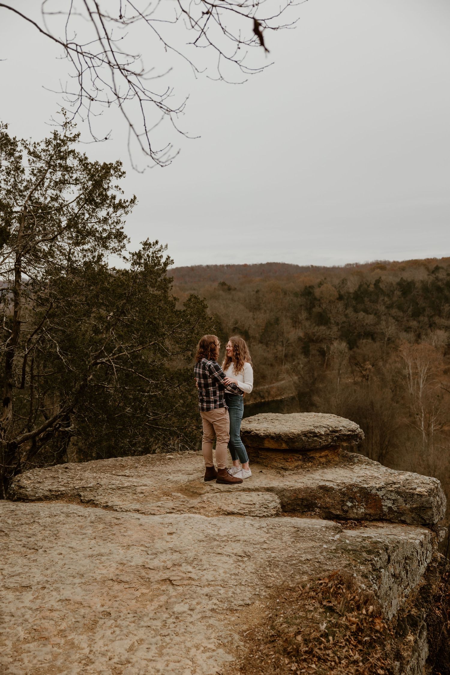 Couple standing close together on a rocky cliff edge with bare trees and mountains in background.