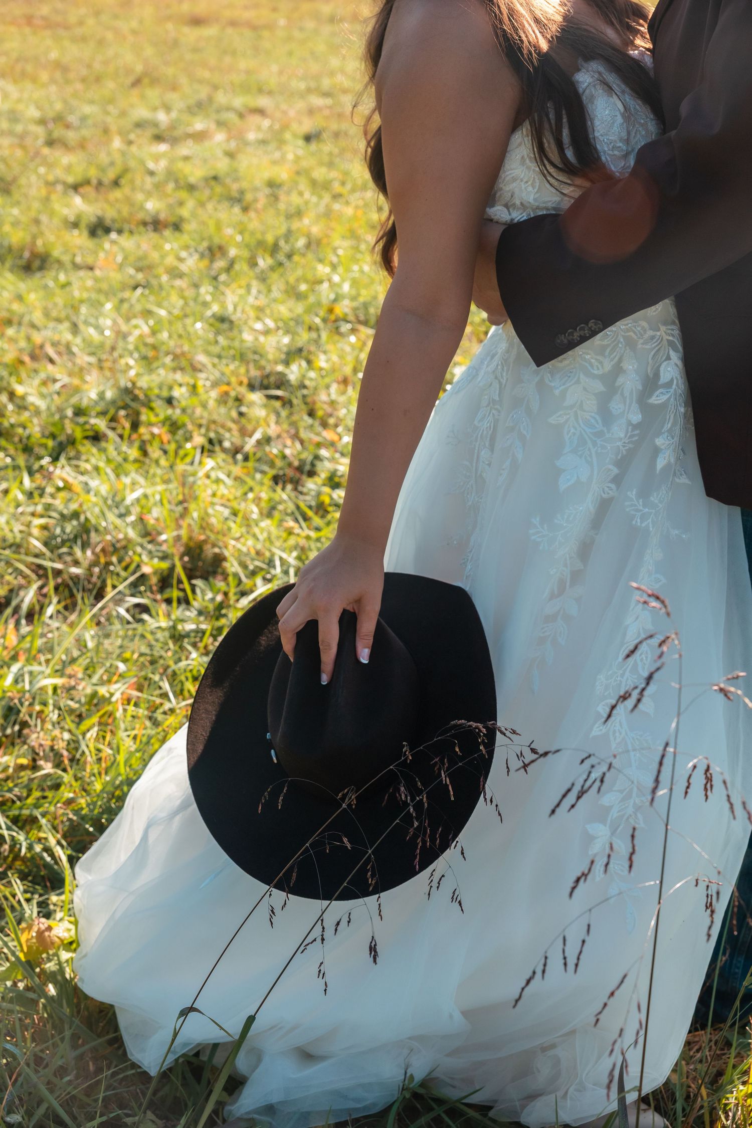 Close-up of a bride in a white lace dress holding a black cowboy hat in a sunlit field.