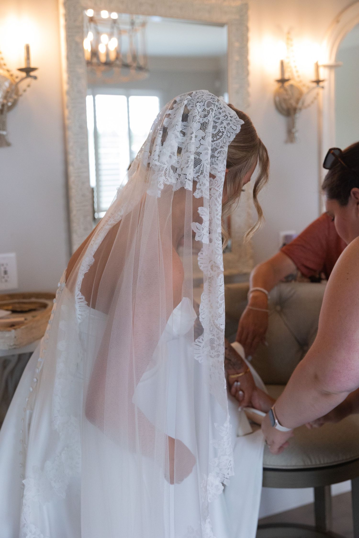 A delicate lace veil being carefully arranged during wedding preparations in an elegant room with wall sconces.