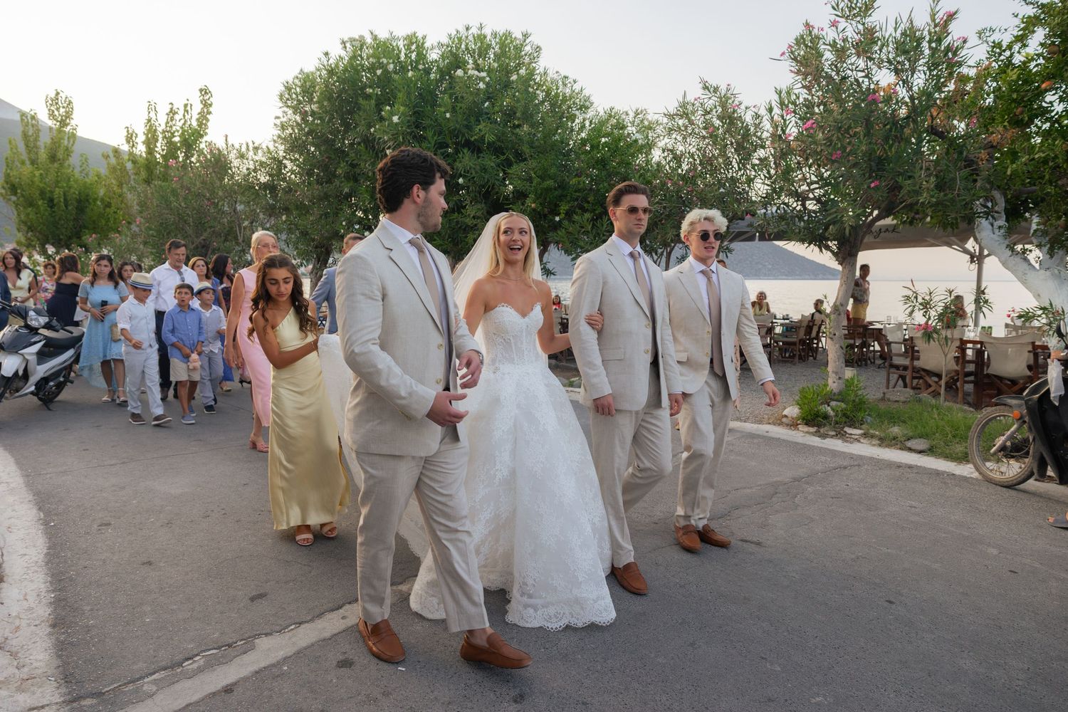 Wedding party in white attire walks along a paved path during an outdoor celebration.