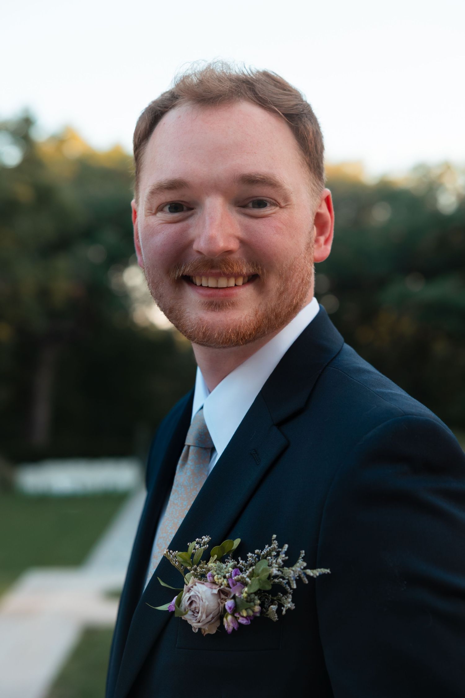 Groom wearing navy suit with floral boutonniere smiles during outdoor wedding portraits.