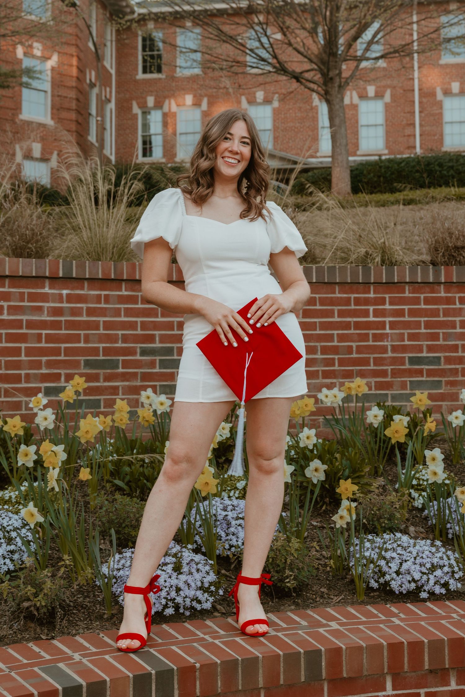 Smiling graduate in white dress holds red cap outside brick building with daffodils blooming.
