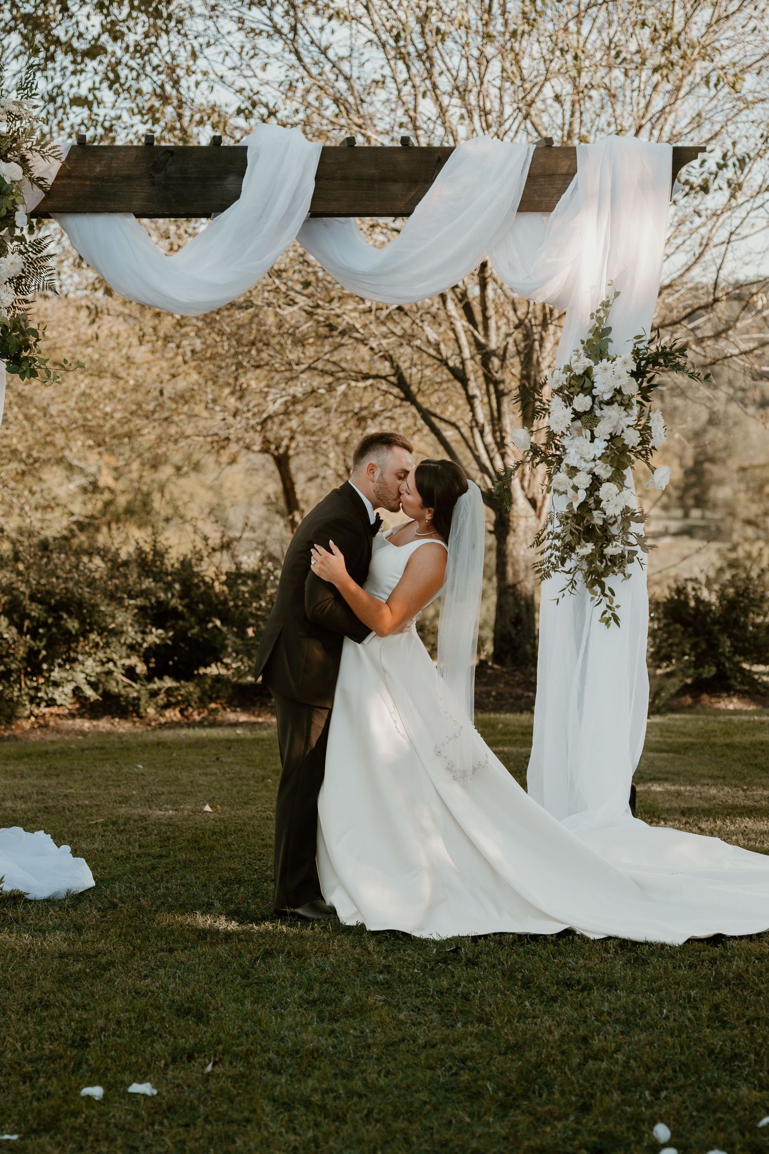 A couple shares their first kiss under an elegant white draped wedding arch with floral decorations.