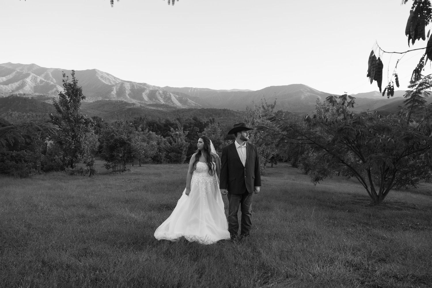 Couple walking through a scenic mountain meadow with rolling hills and dramatic sky in black and white.