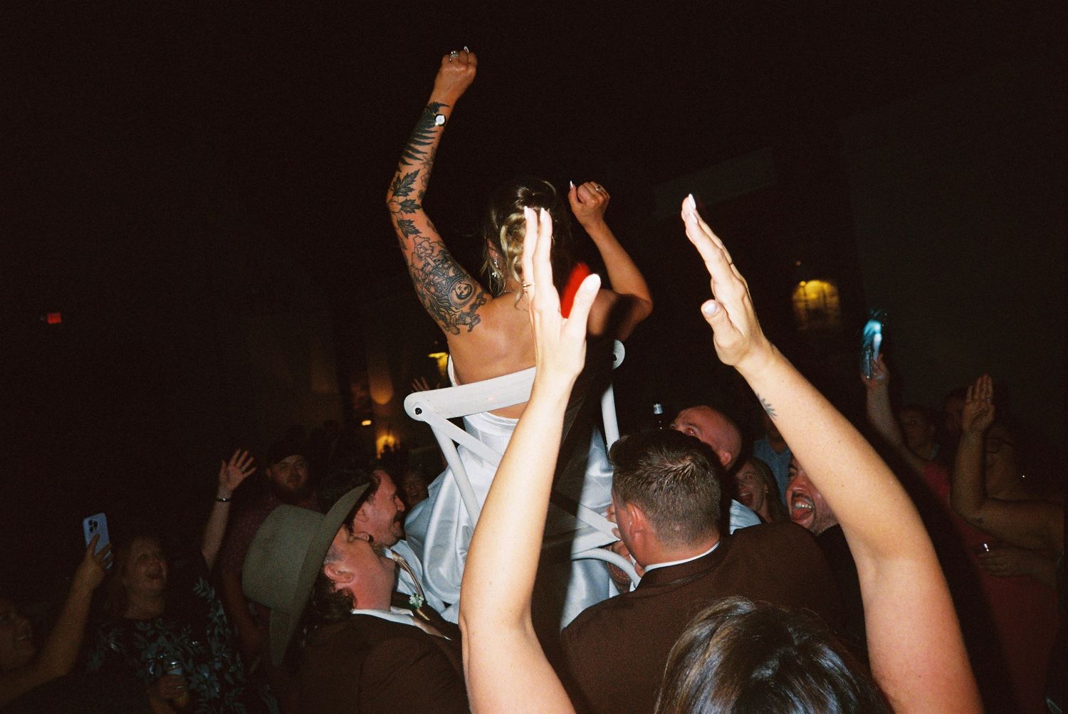 Energetic crowd at a wedding reception with hands raised in celebration on a dark dance floor.