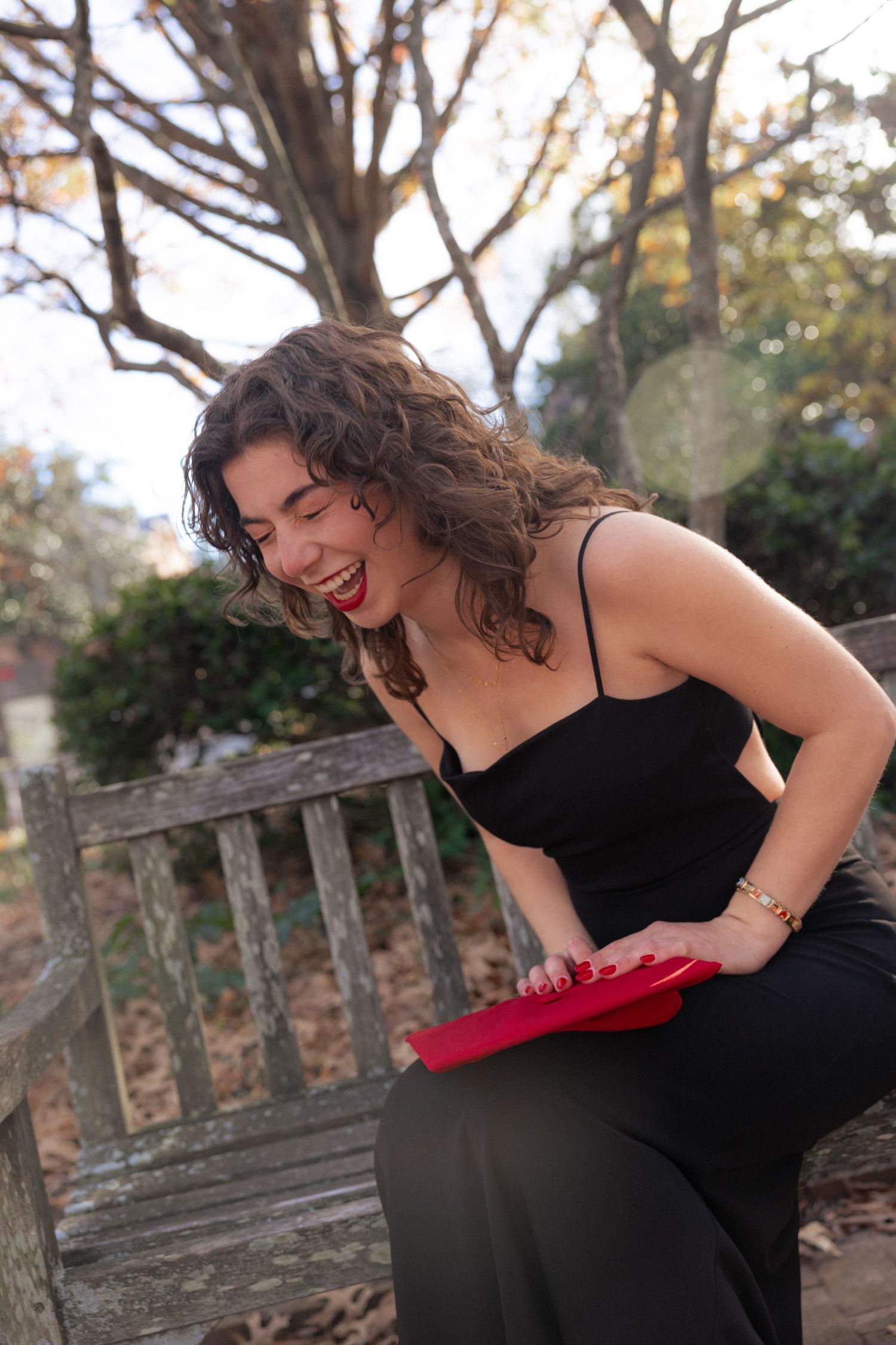 A laughing woman in a black dress sits on a park bench holding a red book, with autumn trees in the background.