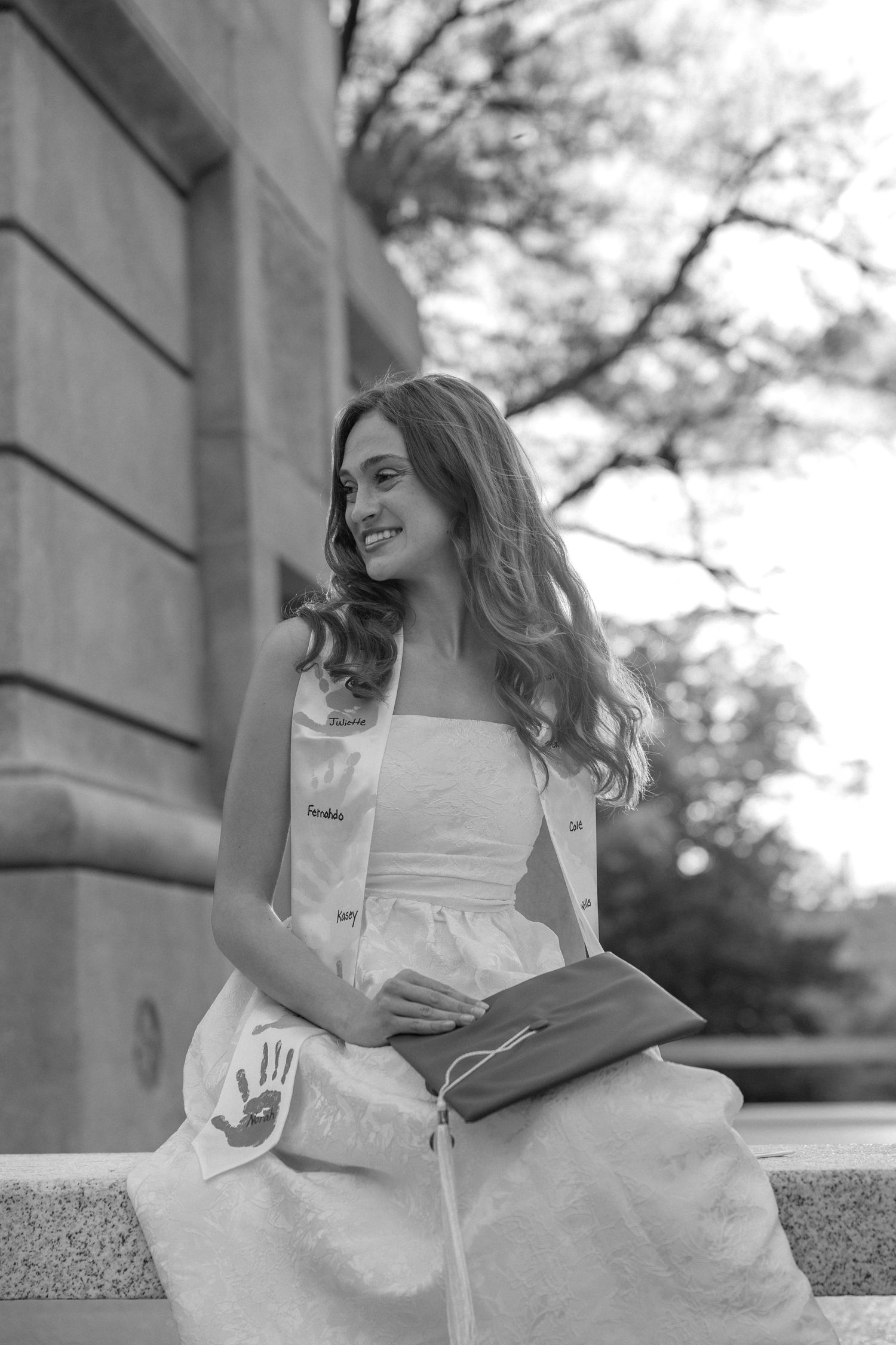 Young woman in white dress and graduation sash sits outdoors smiling, holding diploma in black and white photo.