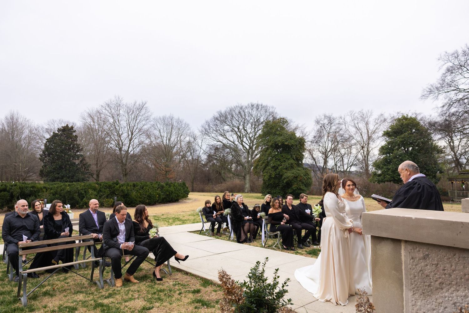 An outdoor wedding ceremony takes place on a stone terrace with guests seated on wooden benches surrounded by winter trees.