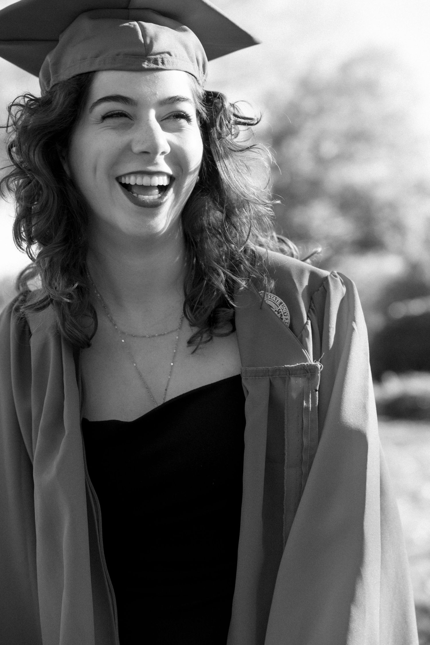 Black and white photo of a young woman laughing in graduation cap and gown outdoors.