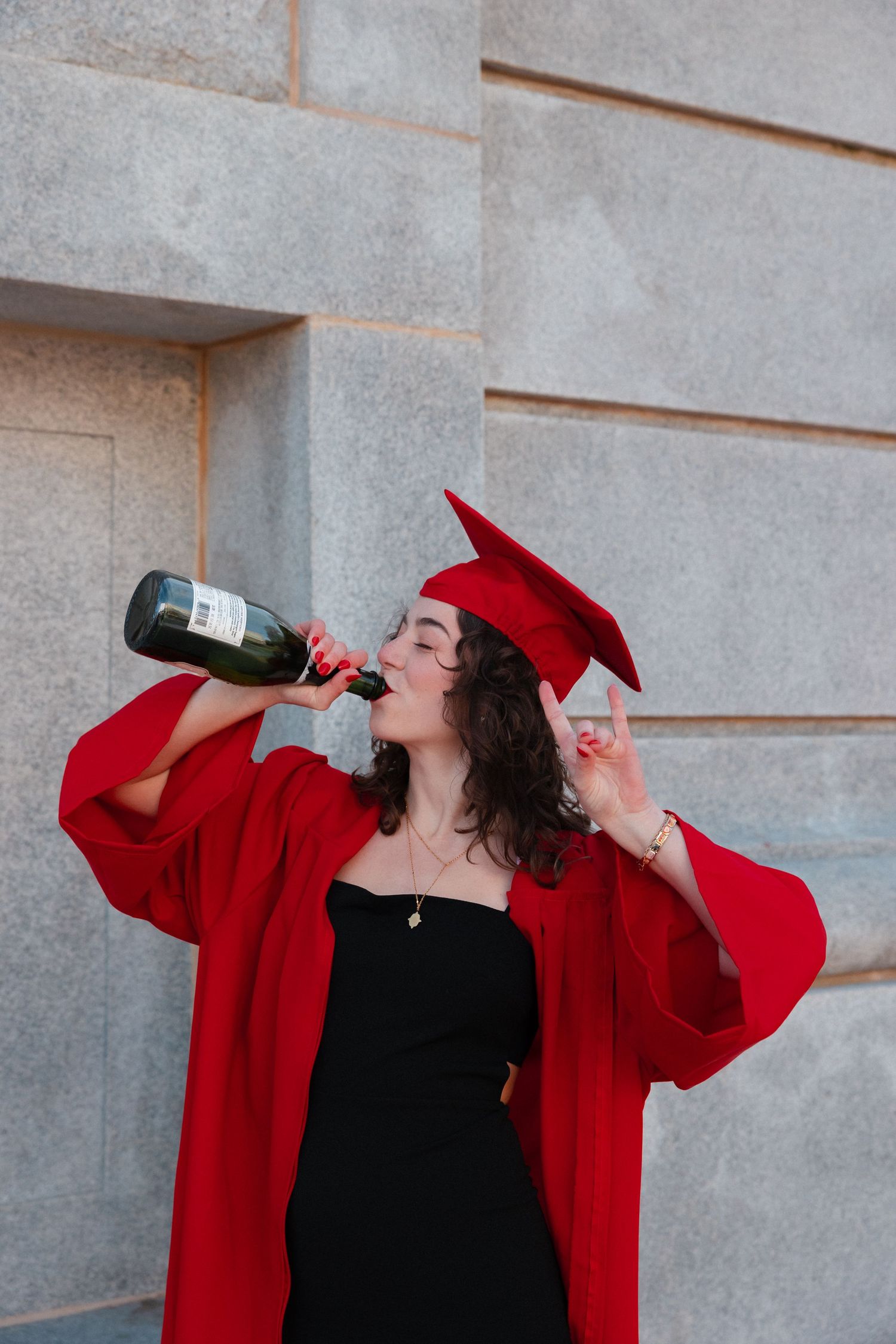 A graduate in red cap and gown celebrates by drinking champagne from a bottle outdoors.
