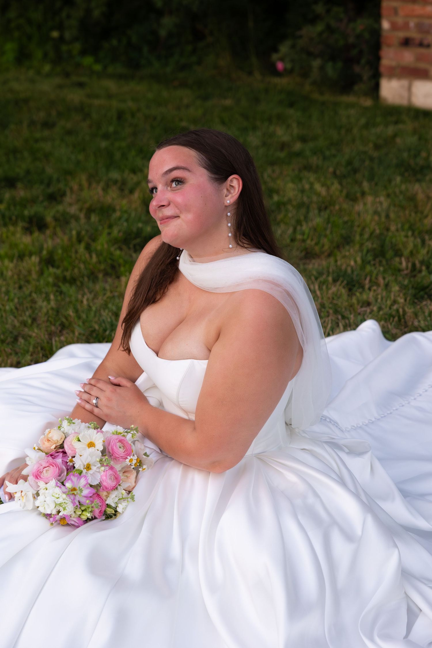 A bride in a white wedding dress poses gracefully on grass with a colorful bouquet of pink and white flowers.