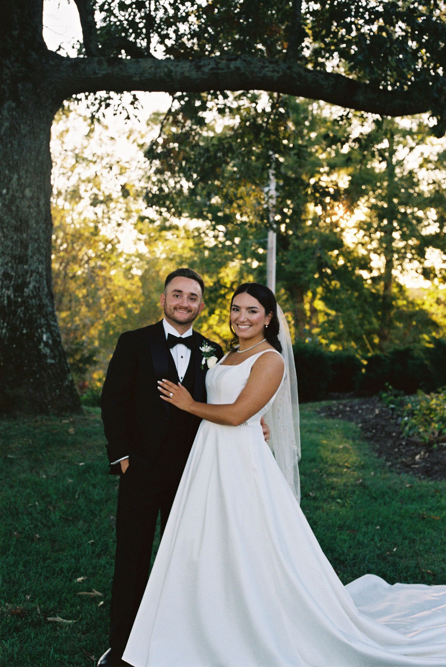 Newlyweds pose together outdoors in formal wedding attire with flowing white dress and black tuxedo.