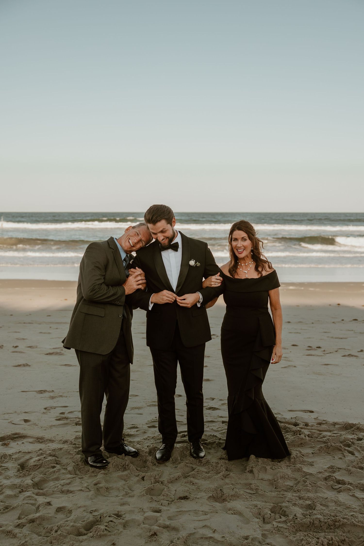 Three elegantly dressed people share a candid moment of laughter on a beach at sunset.
