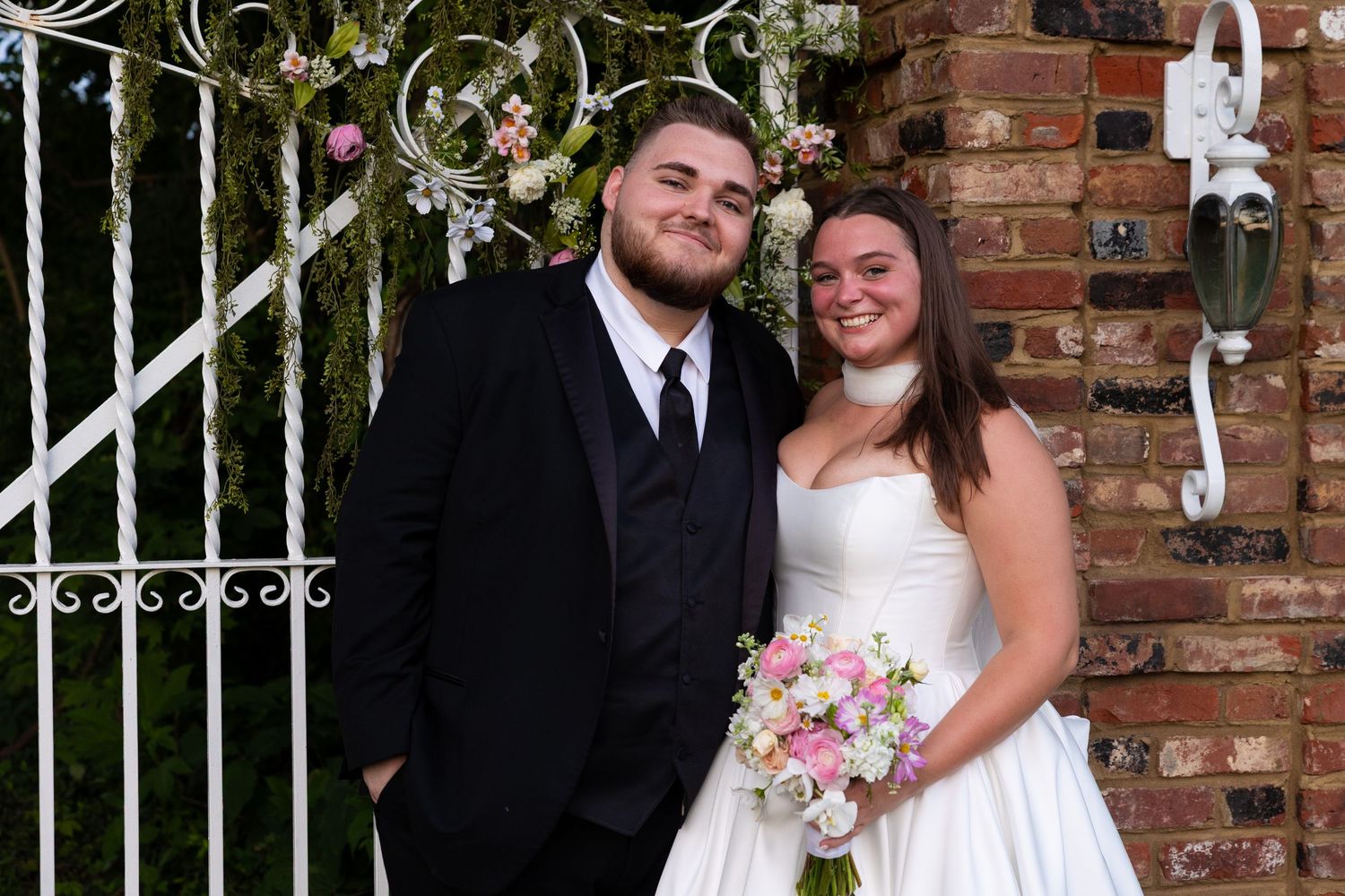 A bride and groom pose together in front of a decorative iron gate and brick wall.