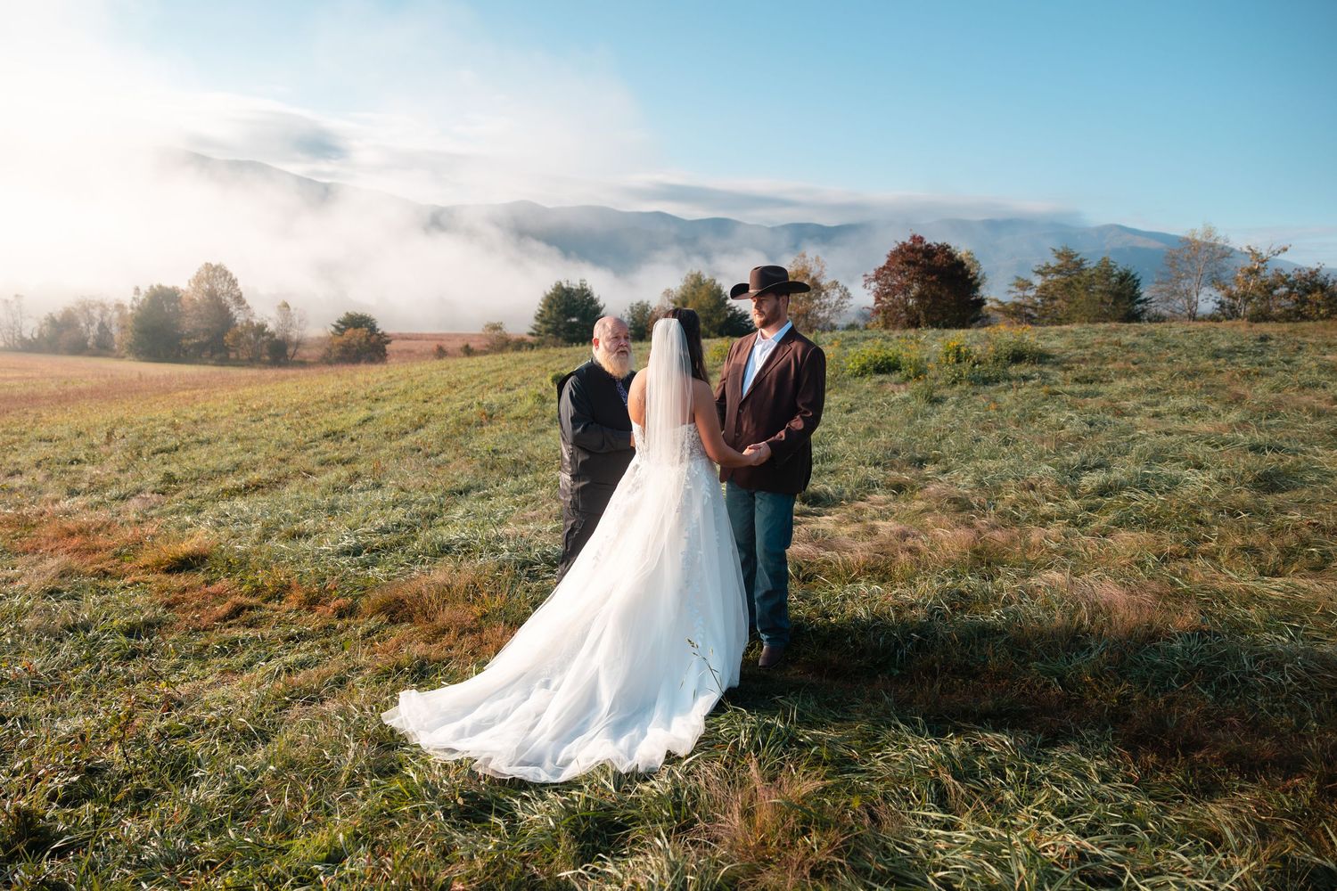 A couple in wedding attire embraces in a sunlit meadow during golden hour with rolling hills in the background.